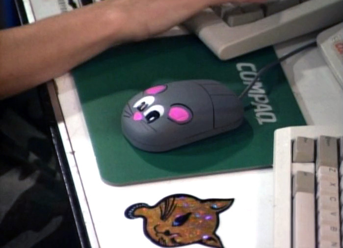 Close-up of Sydney's computer mouse from Superhuman Samurai Syber-Squad. The mouse has stickers on it to make it look like a mouse mouse. It's on a Compaq mousepad that's between two keyboards; a winking cat sticker is also on the desk.