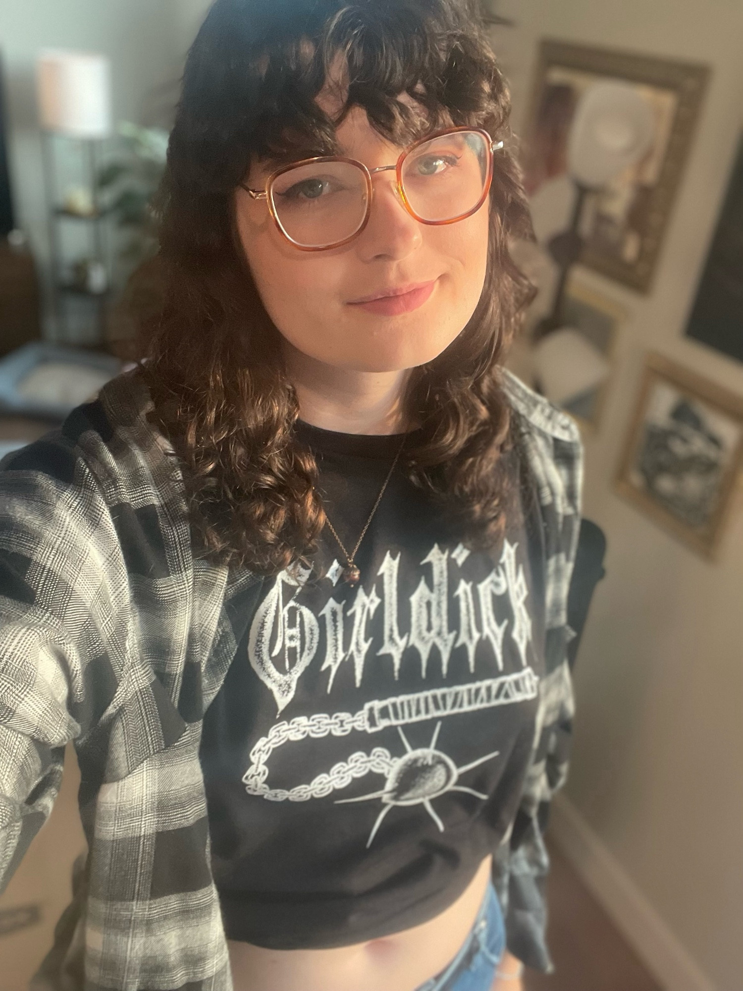 Moonreed smiling into the camera, wearing a black t-shirt with the word “girldick” on it with a flail drawn bellow, she’s wearing a black and white plaid over shirt and jeans as well.