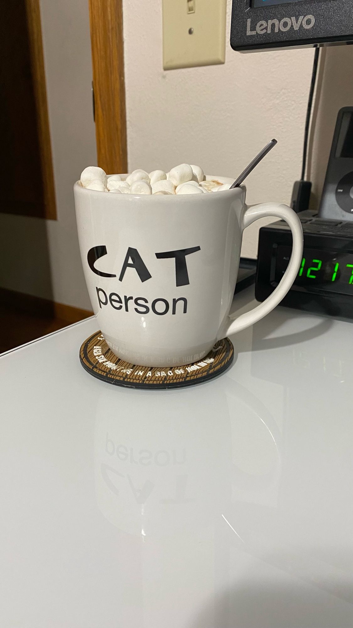 A white mug with text that reads “CAT person” filled with hot cocoa and mini marshmallows rests on a brown coaster on a white desk. The spoon used to stir is still in the mug and can be seen sticking up from a bed of marshmallows on the handle side (right) of the cup.