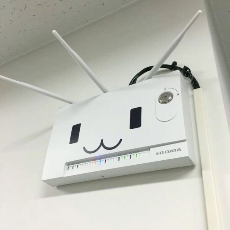 A wall-mounted networking device, likely a wireless router or access point, with three attached antennas. The device has a cute :3 face painted or imprinted on its front side.