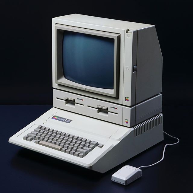 An Apple IIe computer, with an Apple double disk drive and monitor on top of it. The computer has a mouse next to it, connected in the back.