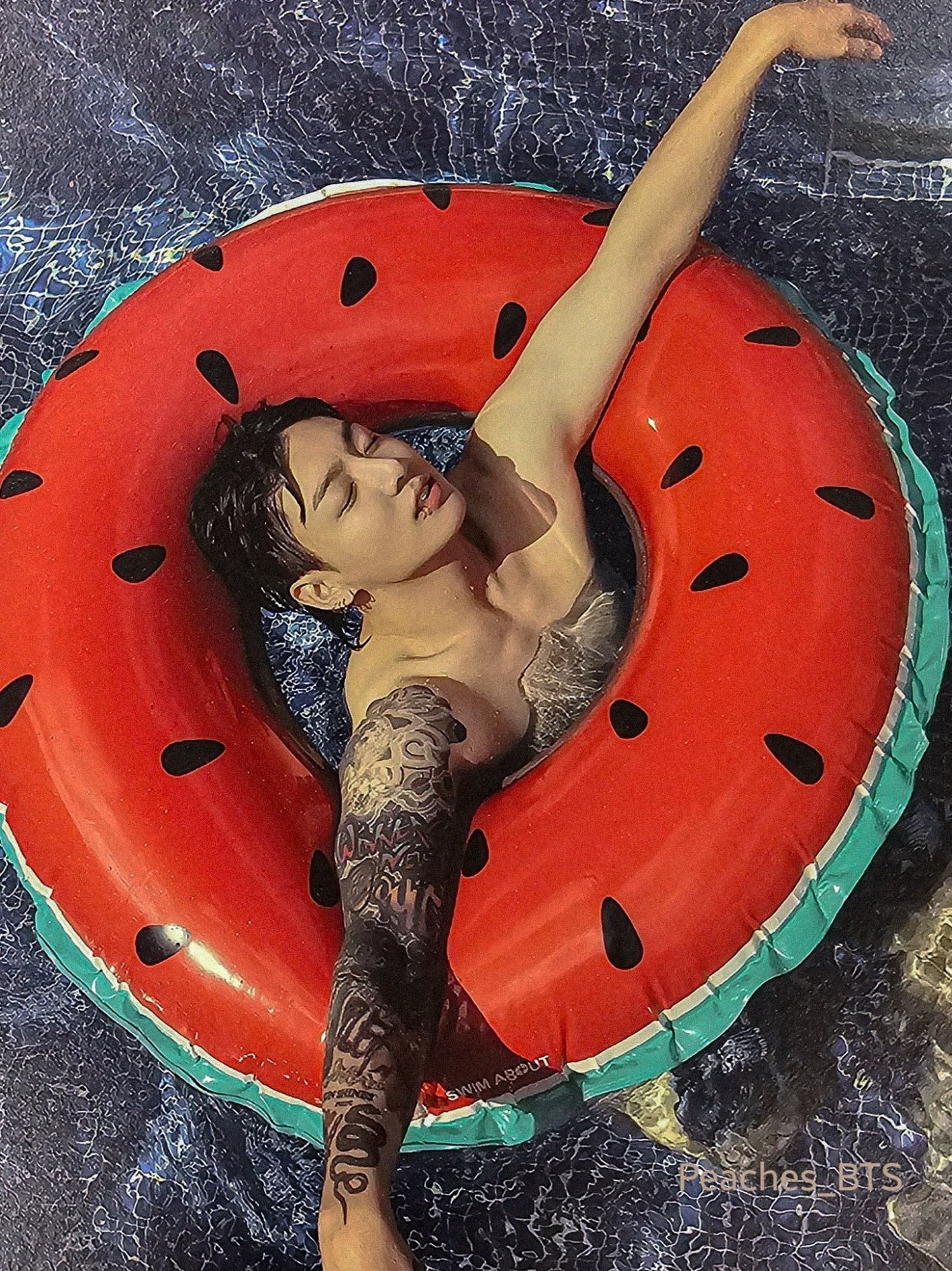 Idol Jeon Jungkook in a pool, he's in a watermelon pool floatie. His eyes are close and his arms are spread out.