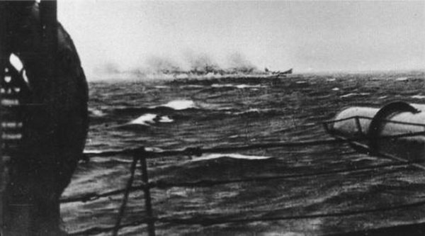 B&W Photo of the battleship Bismarck on fire and heavily damaged taken from the deck of a Royal Navy cruiser preparing to give it the coup de grace with torpedoes.