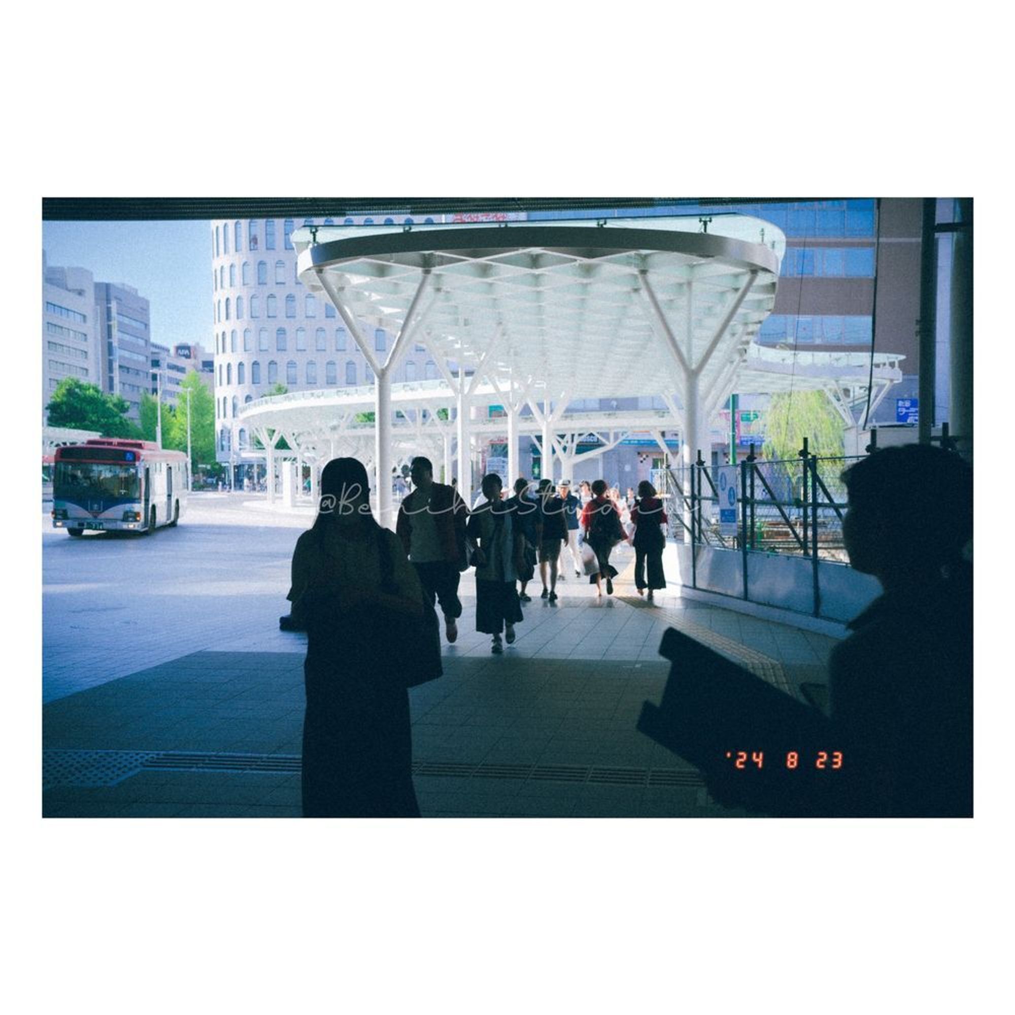 The image appears to be taken at the entrance of a modern bus terminal or station. There is a bright and sunny outdoor scene visible through the large opening, contrasting with the shadows inside the terminal. A group of people, some in shadow, walk towards the exit, silhouetted against the light. In the background, a bus with a red and blue stripe is driving past. The architecture outside includes a distinctive, rounded white structure with large circular columns supporting a canopy. In the distance, there are buildings with many windows, and signs of greenery from trees. 

The image has a vintage, film-like filter that gives it a soft, grainy texture, reminiscent of old photographs. The silhouette of a person on the right side is particularly dark, creating strong contrast with the bright outdoor area. There is also a faint watermark text that reads “@BeiriniStudio” across the center.