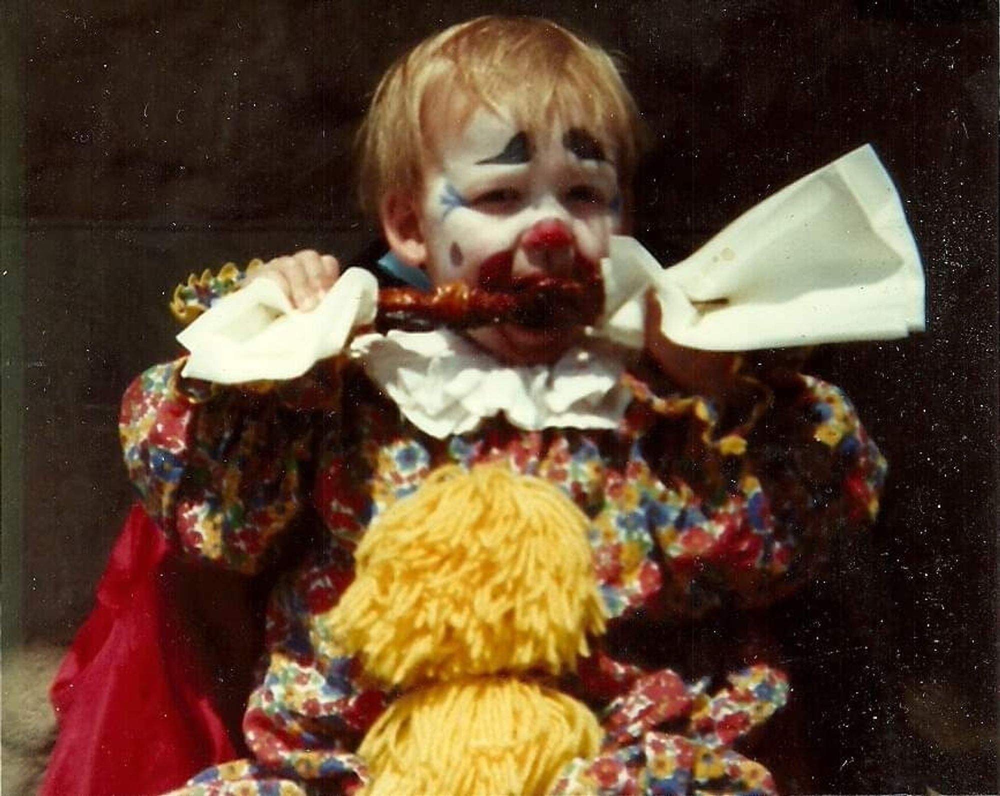 A toddler in full clown regalia and makeup eating sauced meat on a stick