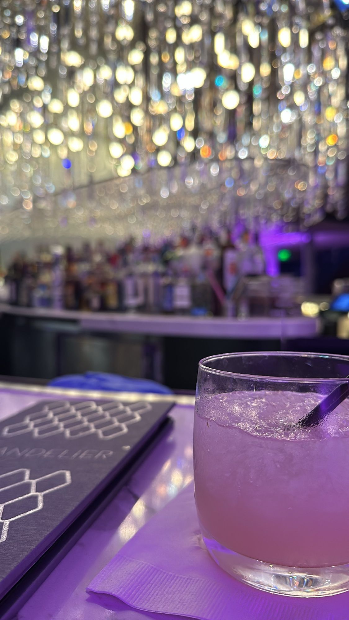 a cocktail in the foreground sitting on a bar with a big crystal chandelier in the background