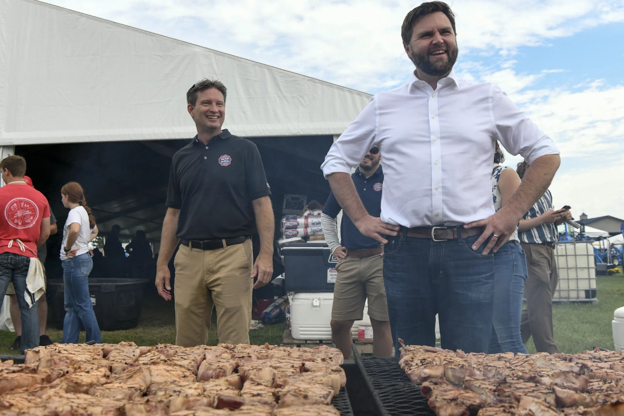 Jd Vance standing in front of a couple of grills that have been piled with chicken (OR  IS IT???)