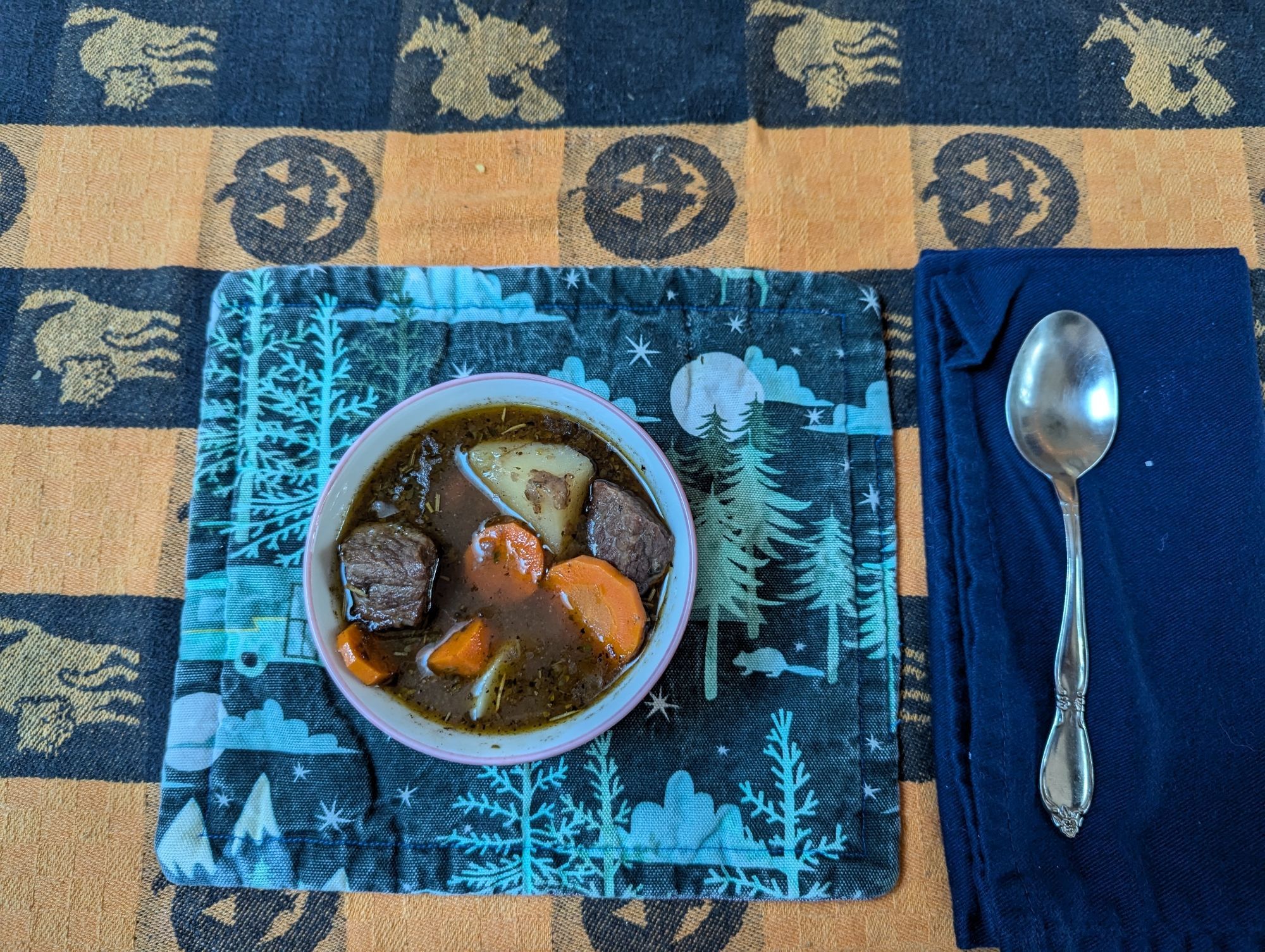 A bowl of stew sits on a placemat with a spoon nearby. The table is covered in a festive pumpkin halloween themed tablecloth.