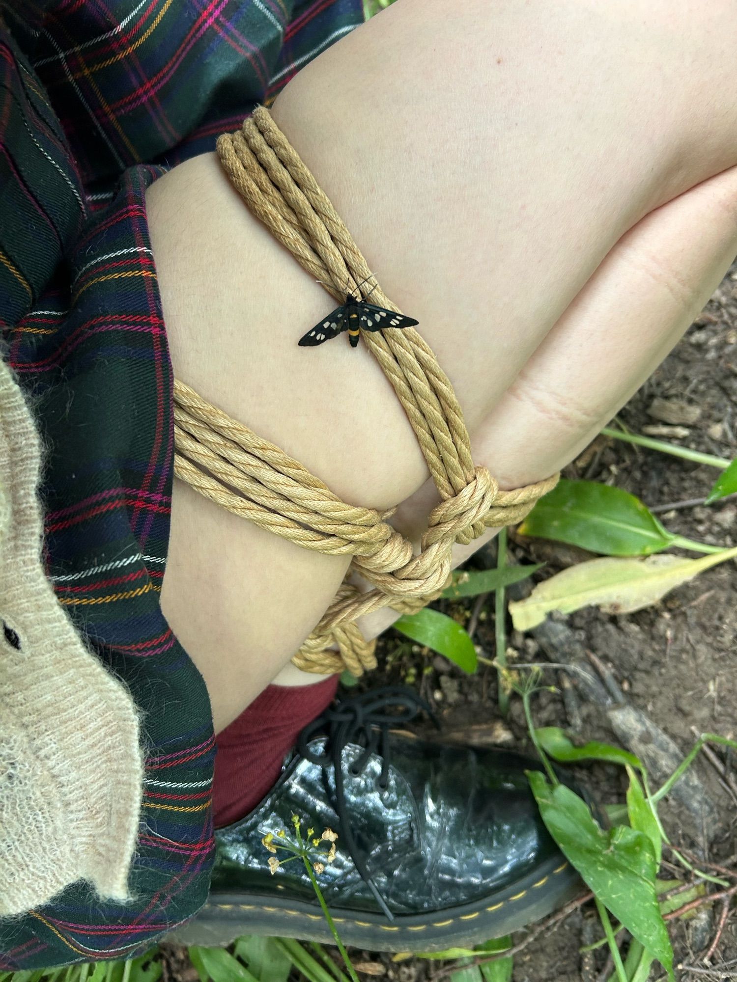 this image shows the leg of a white afab person, sitting in the forest with a self-tied futomomo using jute rope. on it, there is a small black butterfly with white dots