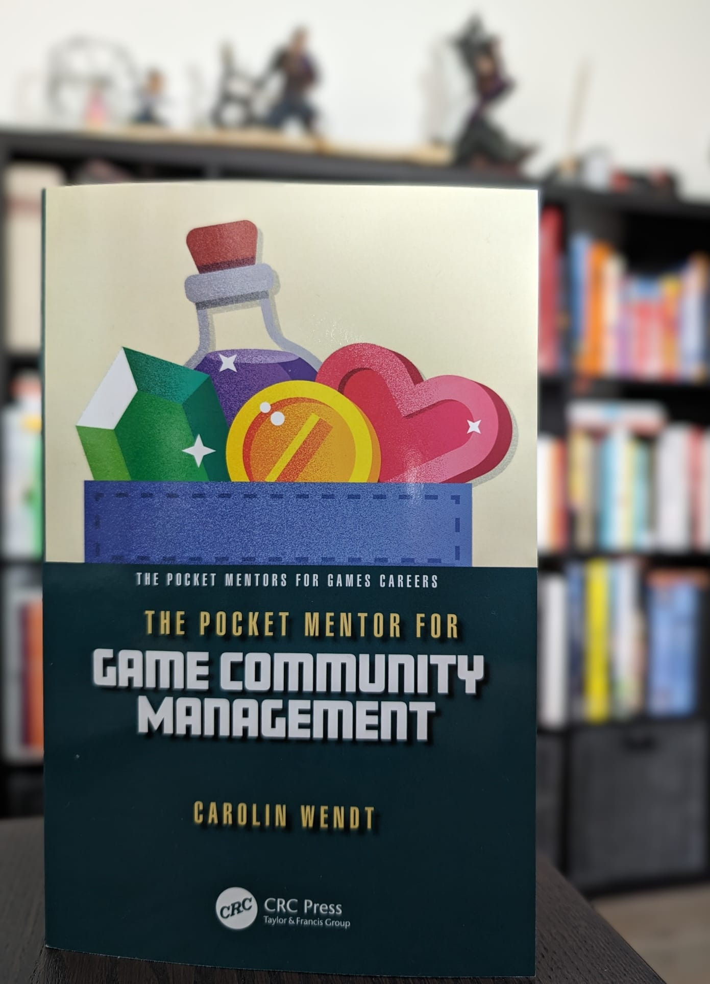 The cover of the book in front of a blurred background with a filled bookshelf. The cover includes a pastel illustration of retro game icons like potions and hearts above the title of the book: The Pocket Mentor for Game Community Management, the author's name and the publisher information.