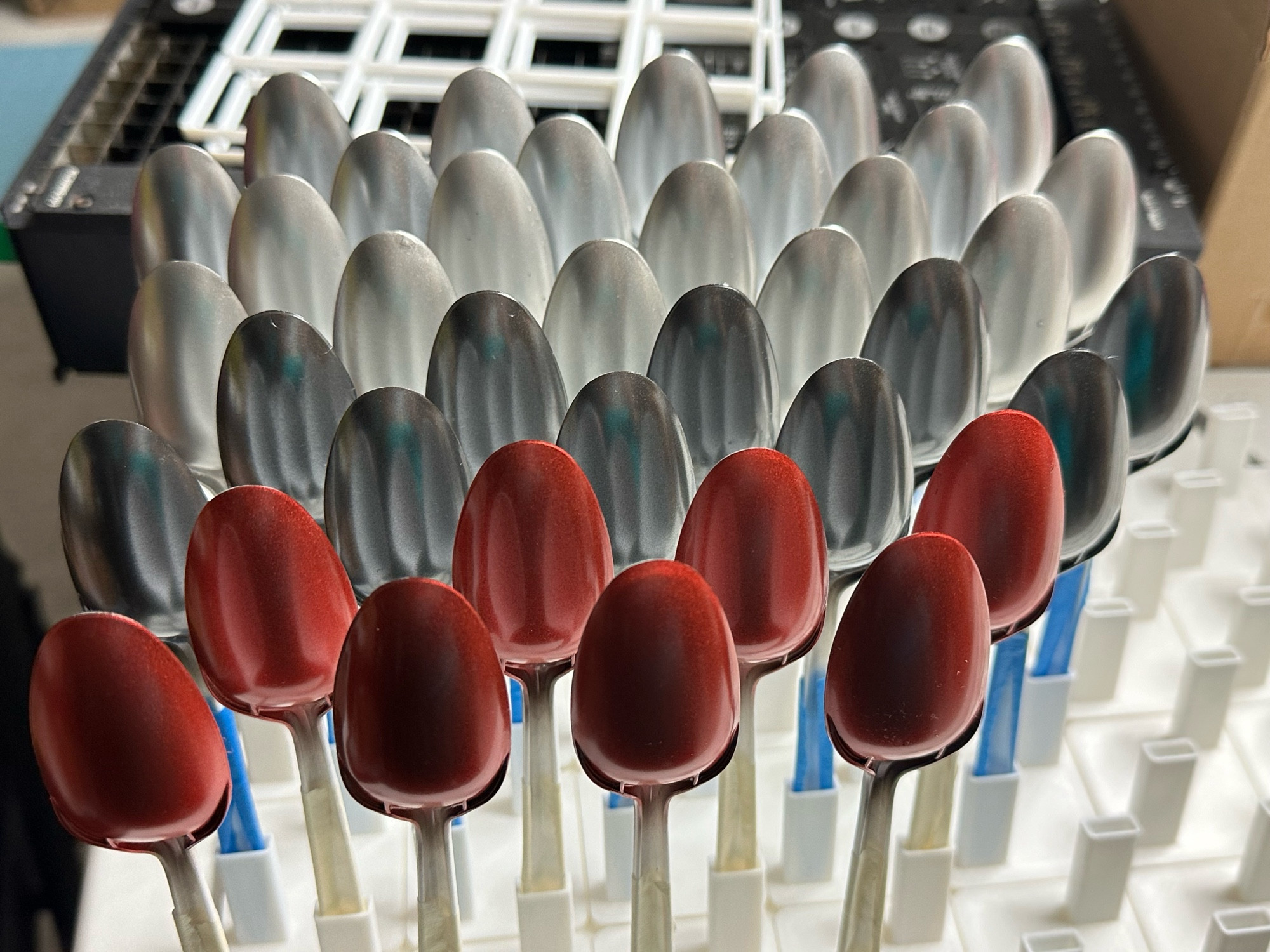 A photo of a few dozen airbrushed plastic spoons held in a carefully designed 3D printed tray, printed for Gridfinity base plates