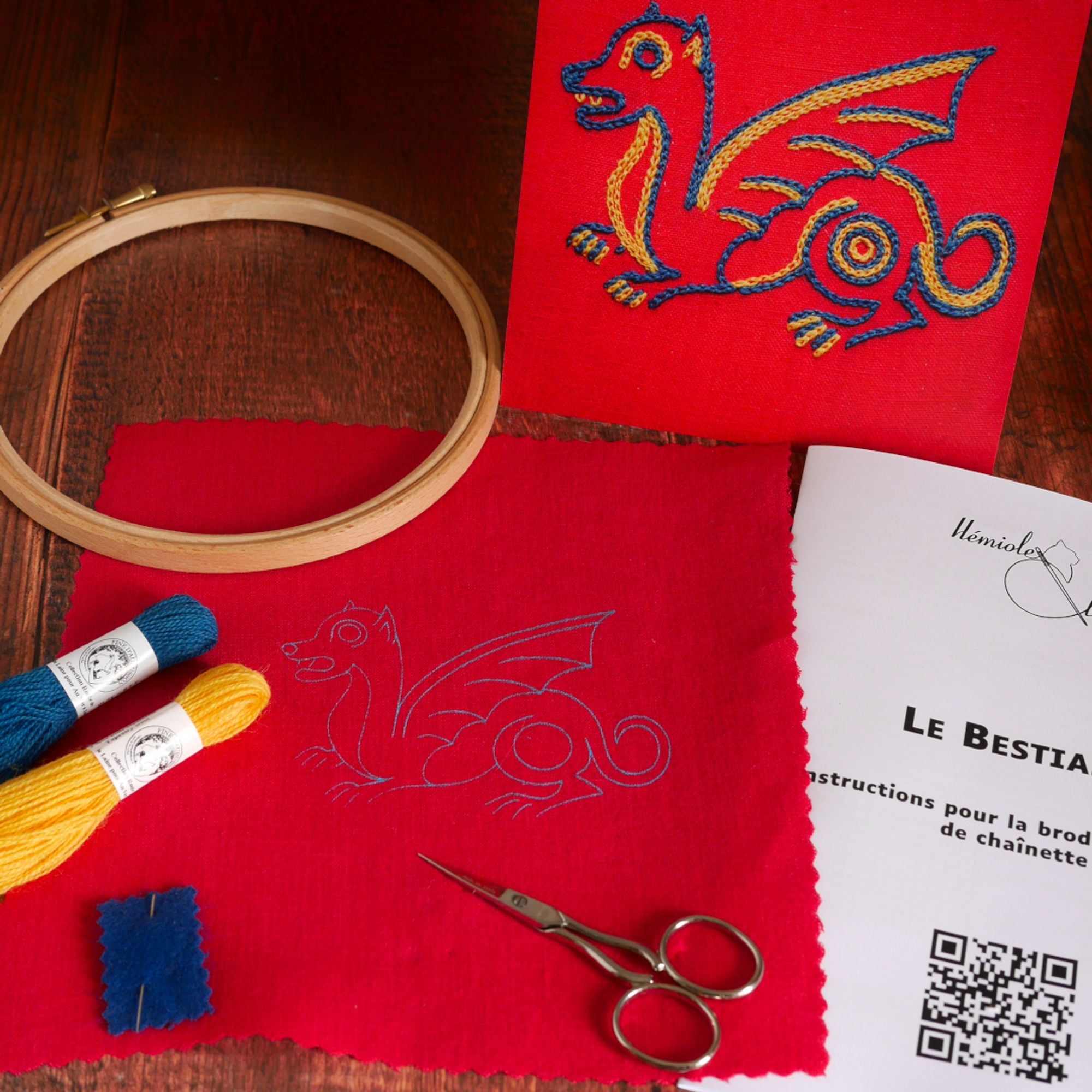 Contenu d'un kit de broderie : la toile imprimée, des fils de laine colorés, une notice, une impression couleur de la broderie finir, un tambour de broderie et une paire de ciseaux de broderie