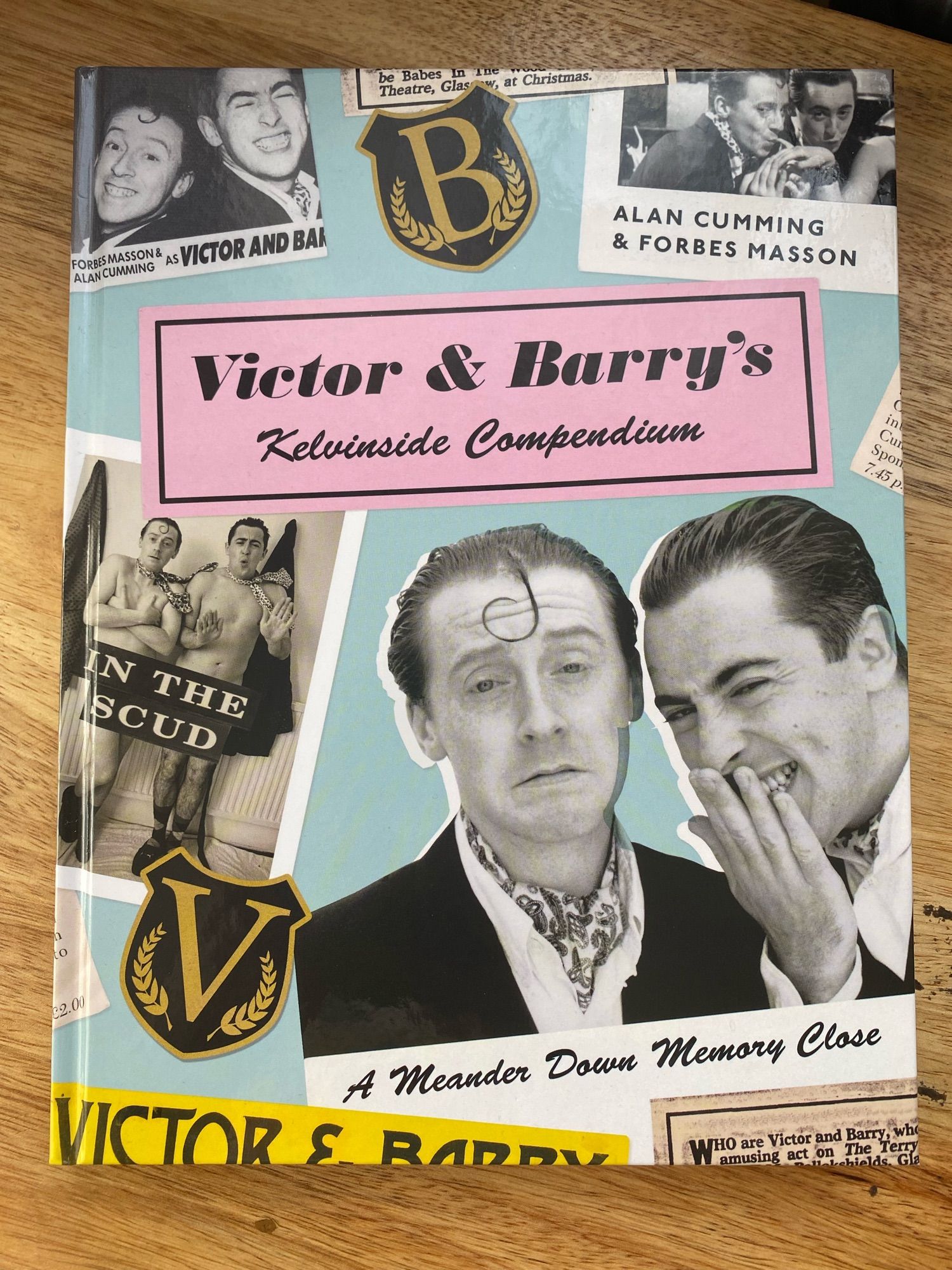 Image of a book on a desk. The book is Victor & Barry’s Kelvinside Compendium: A Meander Down Memory Close. On the cover: Alan Cumming and Forbes Masson.