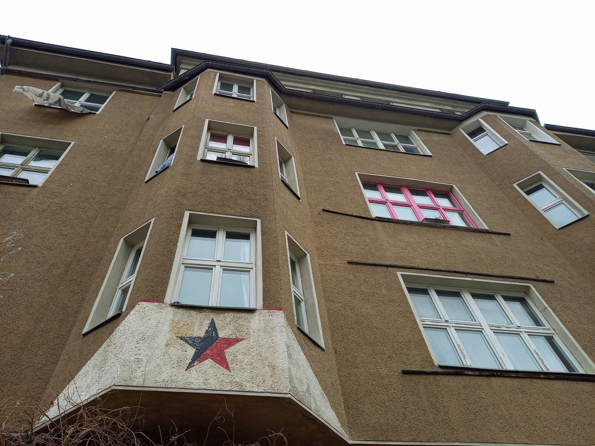 Ansicht der Fassade, großer Altbau, 3 Etagen + Dach, sandfarbender Verputz, große alte Fenster, quadratisch und rechteckig, keine Rundungen, keine Verzierungen, dafür zwei Erker links und rechts vom EG bis zur 3. Etage, über dem Eingang ein rot-schwarzer Stern auf weißem Grund gemalt