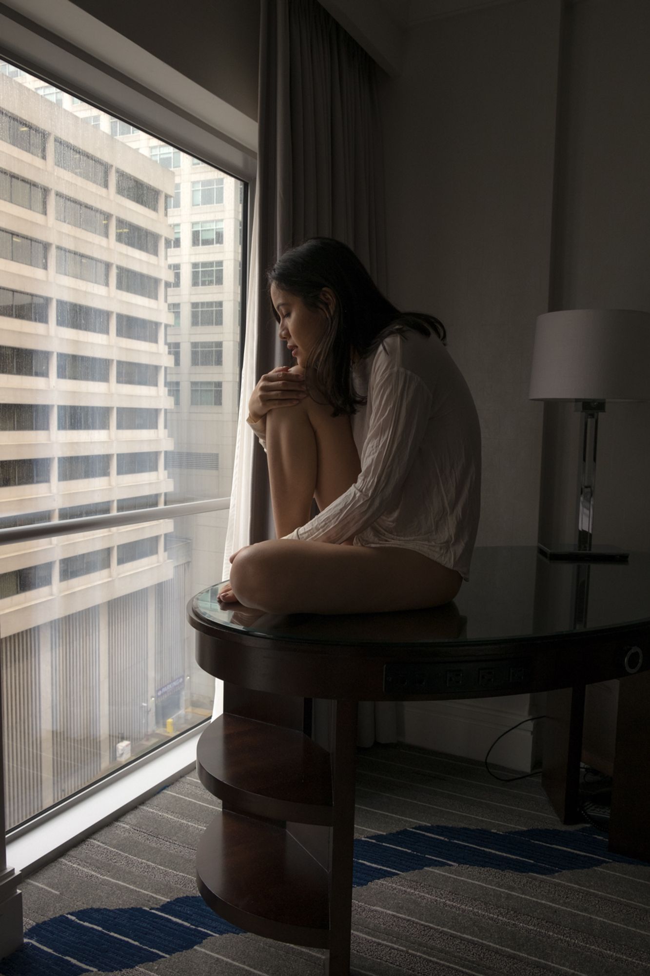 color image of a semi-nude female model wearing a white sheer long-sleeved shirt covering her arms down to her waist sitting on a glass-topped desk looking out a large hotel window one knee pulled up and resting her chin on it with window light highlighting her face and hair