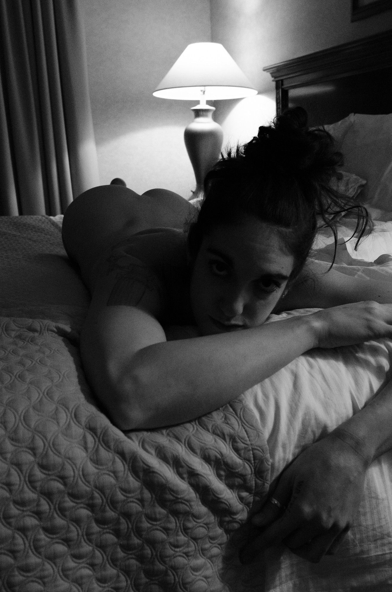 female model lying nude on her stomach on a hotel bed facing the camera with a table lamp in the background leaving most of her body and face in shadow, black hair pulled up in a top-knot