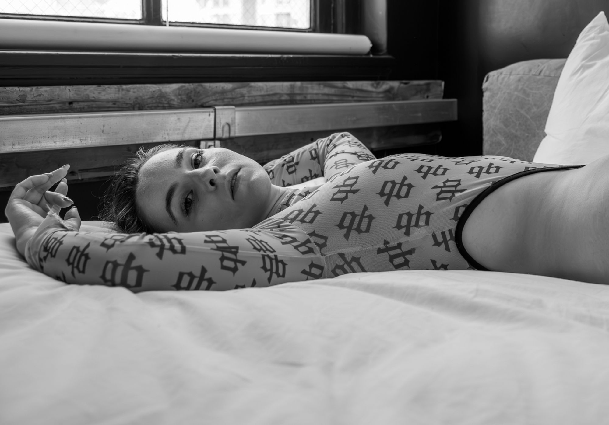 B&W image of a female model wearing a light blue highcut bodysuit covered with the black letter logo 'GB' in a pattern her hip trailing off the right side of the image as she reclines on her back with arm around her head on a bed with white sheets with the bottom of a window visible in the background