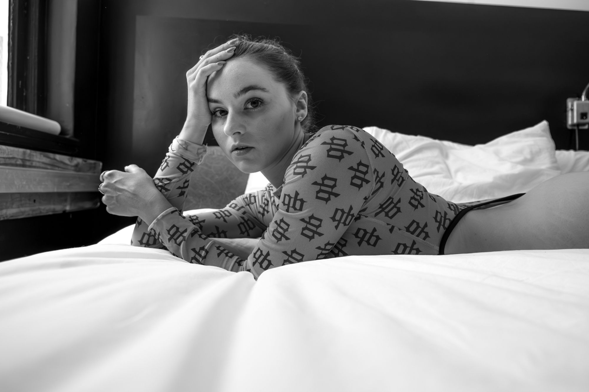 B&W image of a female model wearing a light blue highcut bodysuit covered with the black letter logo 'GB' in a pattern her hip trailing off the right side of the image as she reclines on a bed with white sheets with her head propped on one elbow