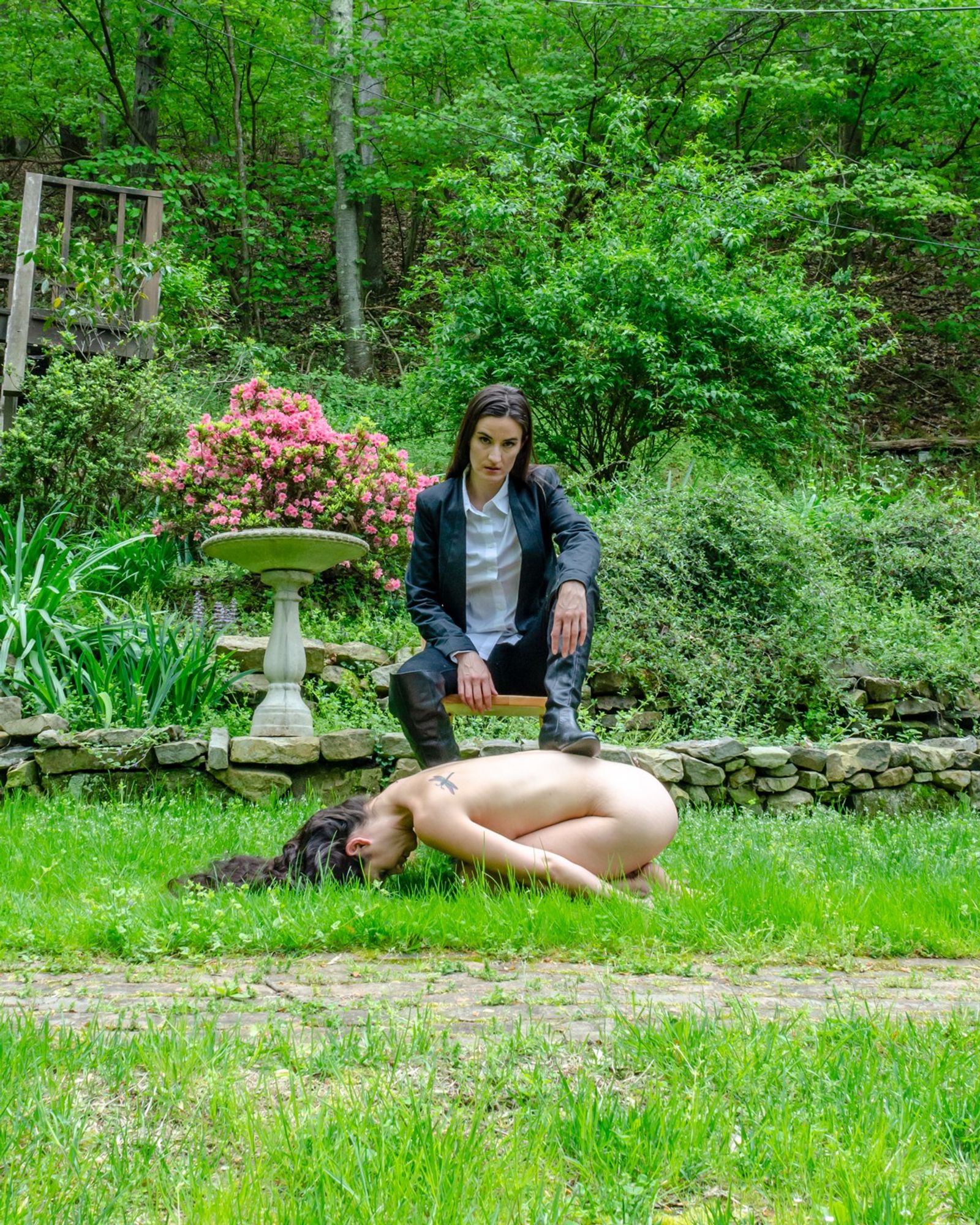 two female models one full nude and kneeling on grass with her head bent down and touching the ground, arms pointing back to her ankles the other model sitting in a chair next to a bird bath dressed in navy pants and jacket with a white shirt and knee-high black boots, one foot resting on the back of the kneeling model surrounded by green vegetation and one pink azalea in full bloom