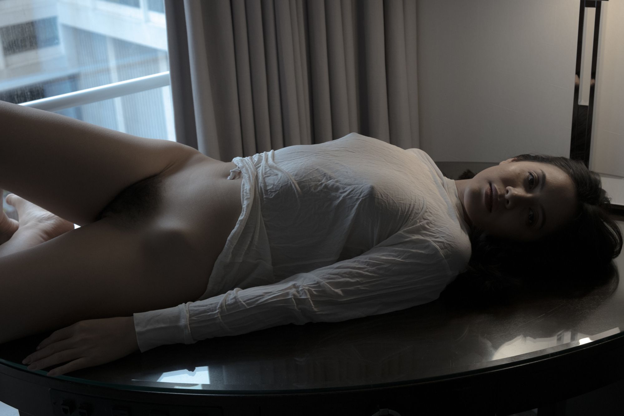 color image of a semi-nude female model wearing a white sheer long-sleeved shirt covering her arms down to her stomach laying on a glass-topped desk looking at the lens with light from a floor lamp highlighting her head and torso her legs shaded by light from a window in the background