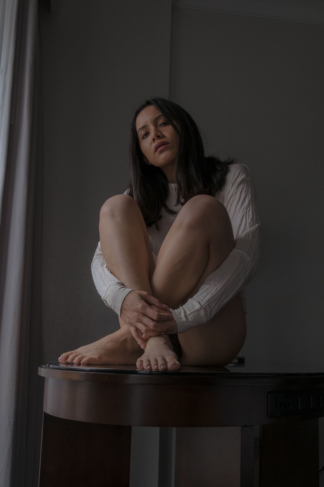 color image of a semi-nude female model wearing a white sheer long-sleeved shirt lit from the side by a window not visible in the image covering her arms down to her waist sitting on a glass-topped desk looking at the lens both knees pulled up with her hands holding her crossed ankles