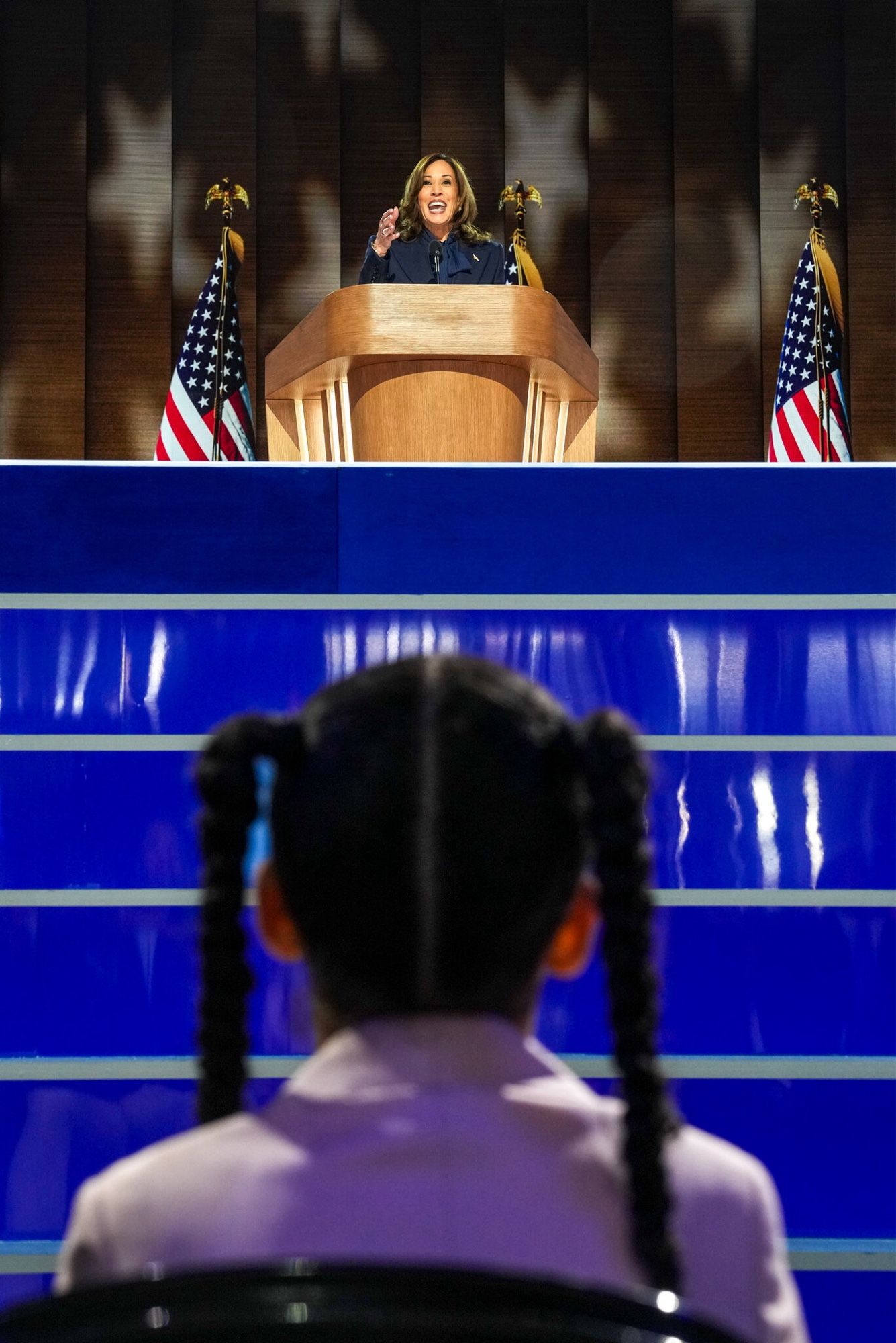 Kamala Harris on the stage at the united center. In front of and below her is the back of a young girl with pigtails’ head.