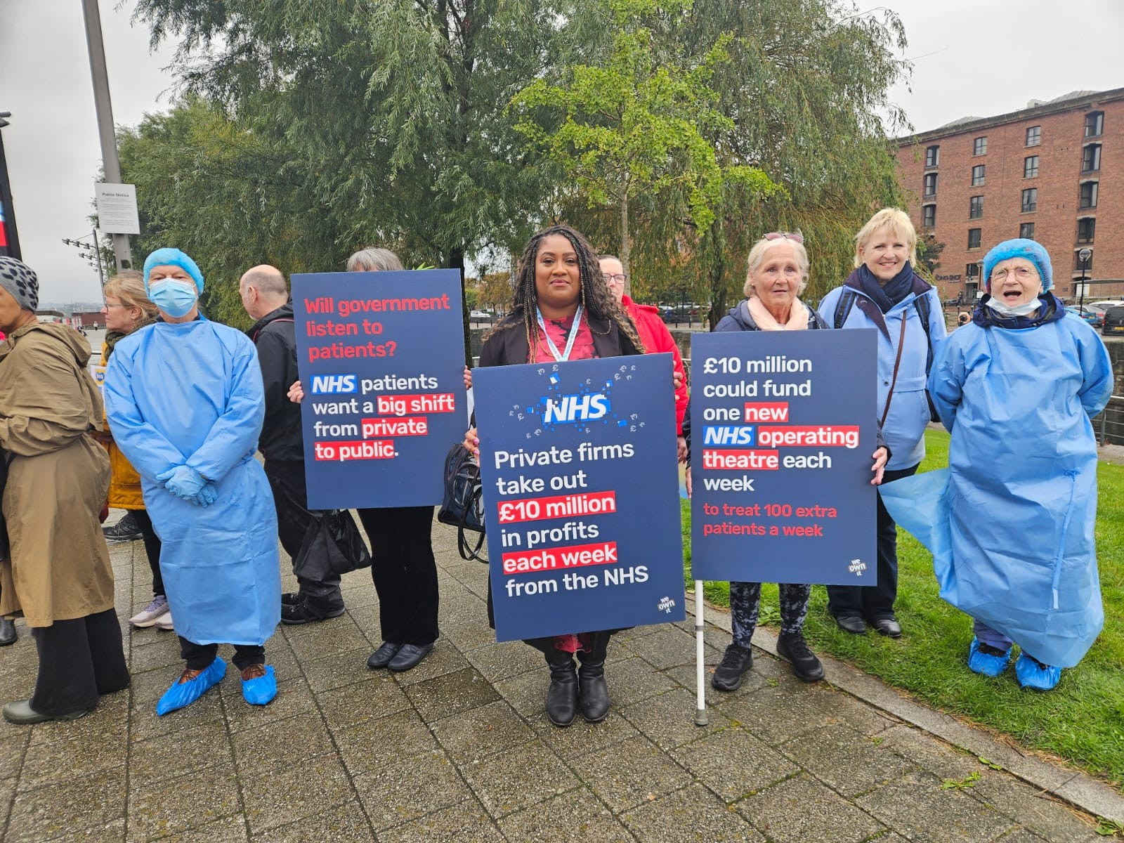 Bell stands with activists in doctor's scrubs. They hold signs highlighting the impact of outsourcing policies on our NHS. Bell's placard says 'NHS: Private firms take out £10 million in profits each week from the NHS'.