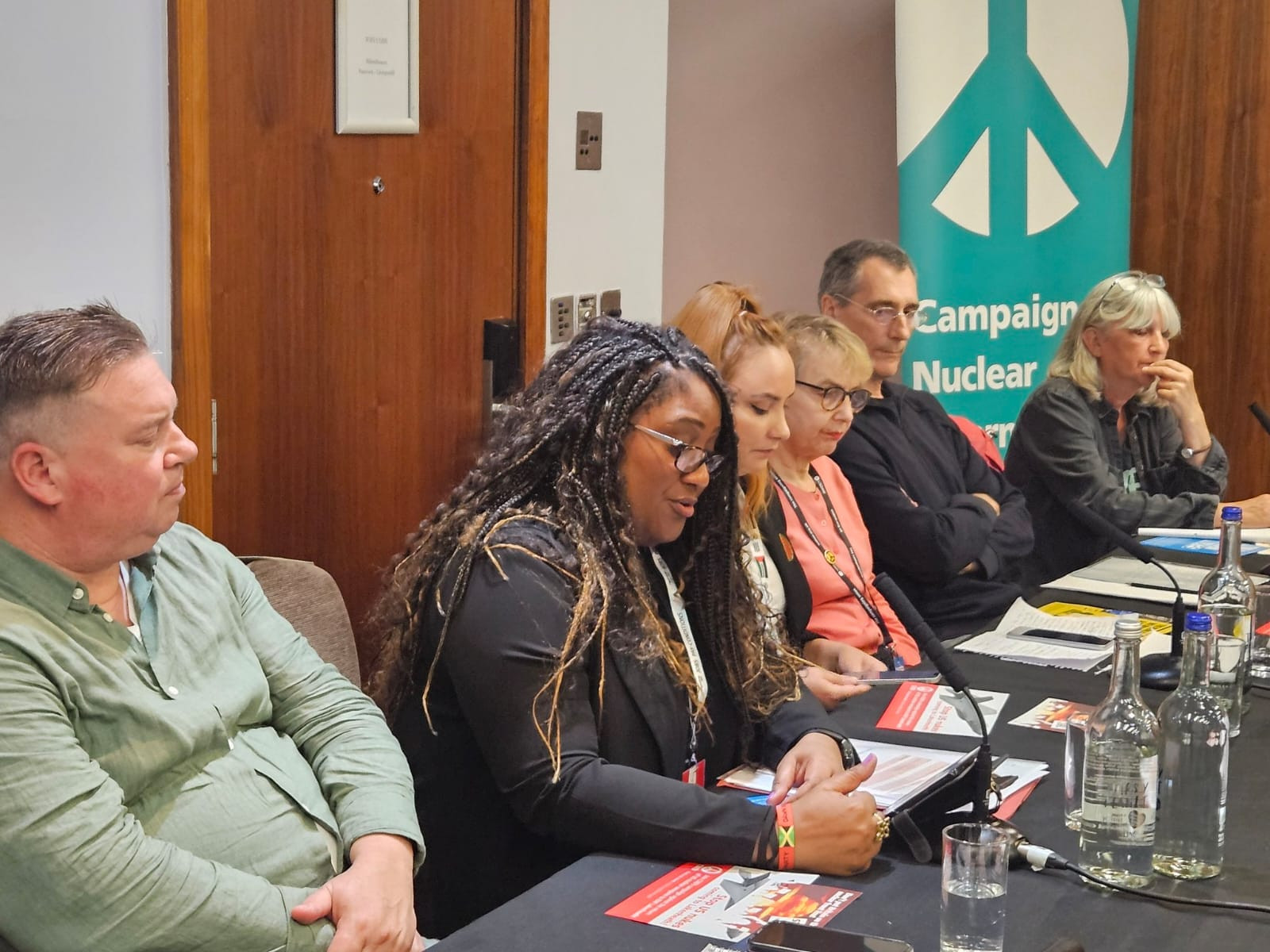 Bell speaks into a microphone on a panel with five other people. There are leaflets and glasses of water arrayed in front of them and a Campaign for Nuclear Disarmament banner in the background.