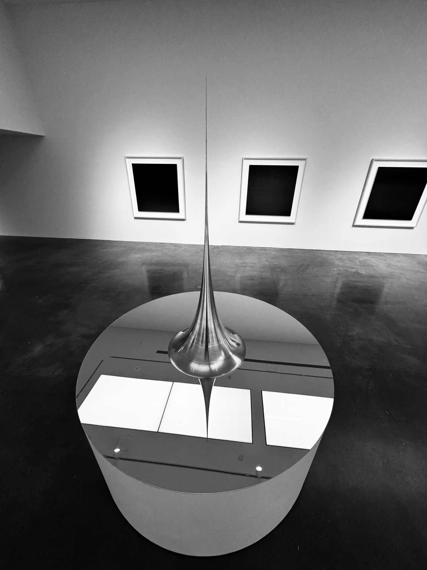 GPT-4o: This image showcases a sleek, modern art installation featuring a tall, spire-like sculpture positioned on a reflective circular surface. The sculpture’s needle-like design extends vertically from the center of the reflective base, creating a mirrored illusion that emphasizes symmetry. The surrounding space appears to be a minimalist gallery with clean lines, polished floors, and plain walls adorned with three framed, black, rectangular artworks in the background. The monochromatic black-and-white composition adds to the contemporary and abstract atmosphere of the scene.