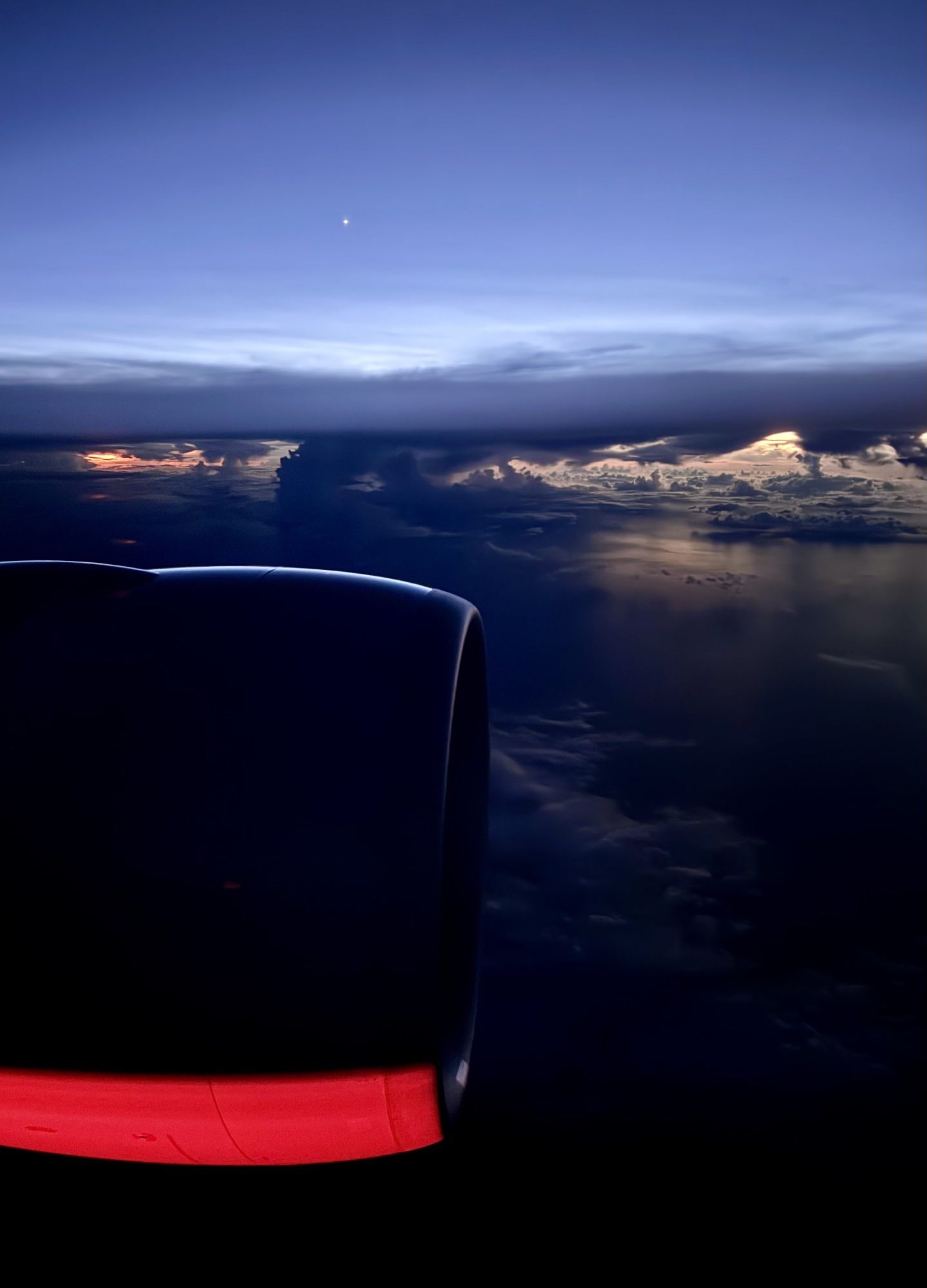 GPT-4o (edited): The image shows a view over an ocean from the window of an airplane, featuring the aircraft’s engine in the foreground. The sky above is transitioning from dusk with a deep blue hue, and there is a clear view of a small, bright planet visible in the upper center of the sky. Below, dark clouds are visible, with hints of light from the setting sun illuminating parts of the horizon and reflecting from the ocean's surface. The airplane engine has a red glow on its underside from the aircraft’s lighting. The scene captures a peaceful and serene moment above the clouds.