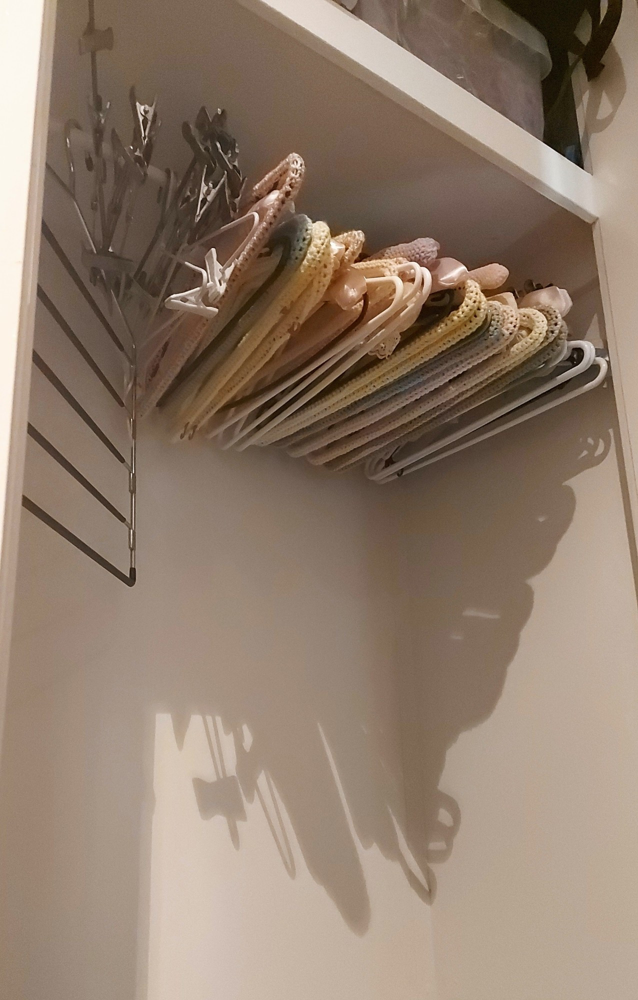 A photo of the inside of a closet. All the clothes have been removed but a large number of empty hangers remain. Most have crochet or sewn covers in soft pastel colours