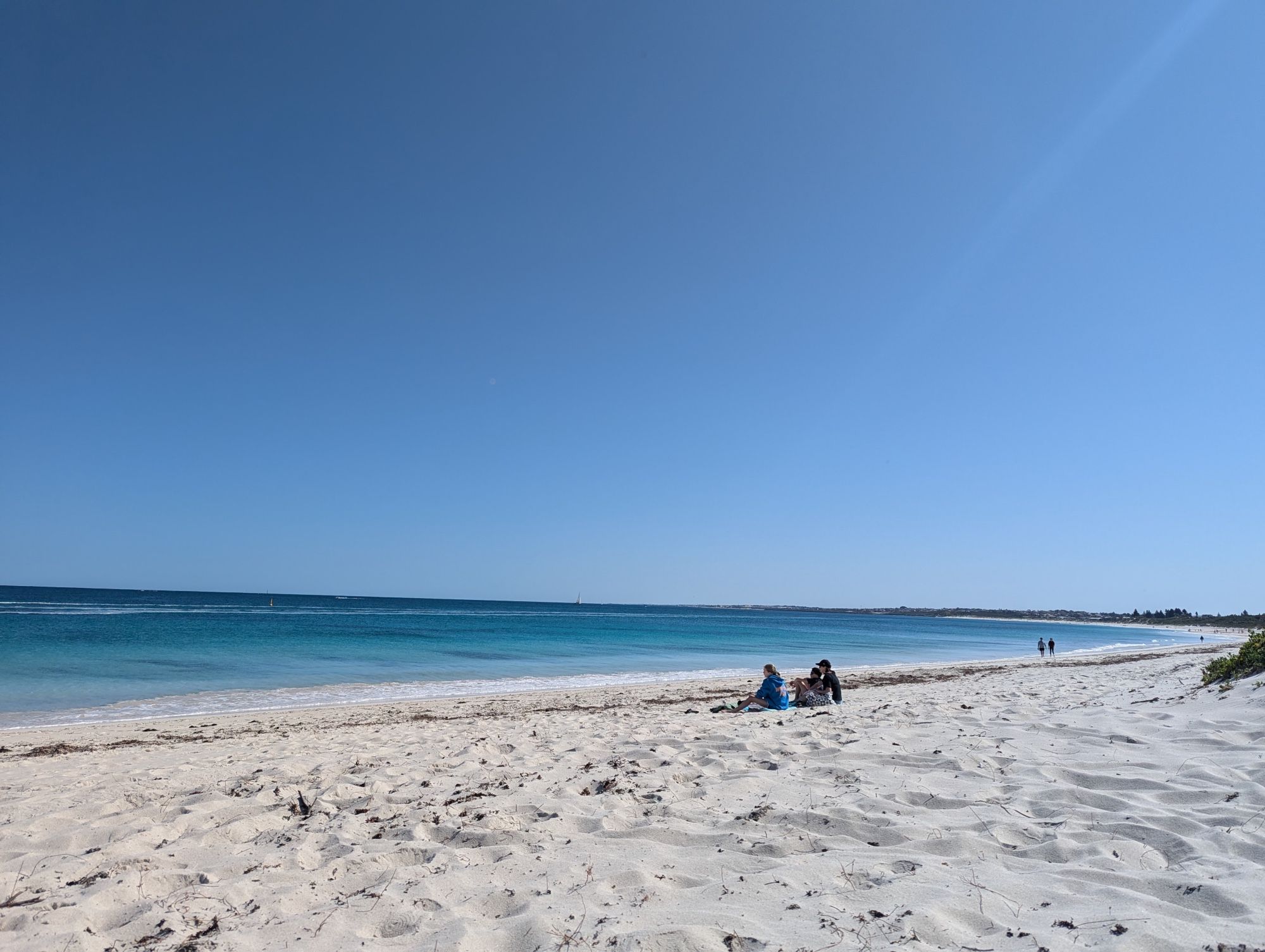 Ein wolkenloser Himmel, ruhige tiefblaue See, weißer Strand, ein paar Menschen sitzen , weiter weg sind 2 Spaziergänger, ganz hinter sieht man kleine Punkte die wohl Menschen sind