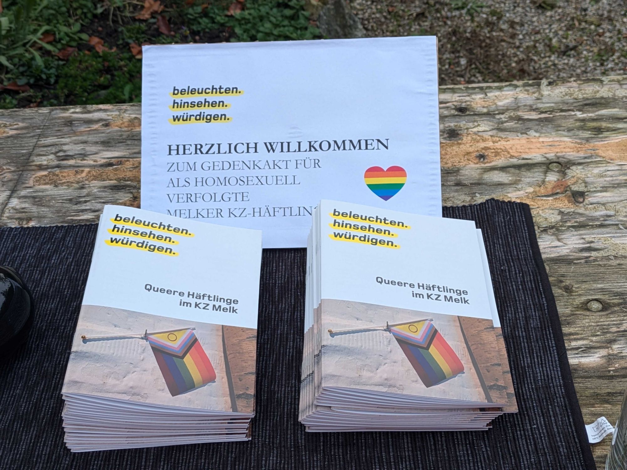 Tisch im Freien, auf dem Broschüre des Projektes "beleuchten - hinsehen - würdigen" zum Thema Queere Häftlinge im KZ Melk liegt. Ein Schild heißt alle herzlich willkommen.