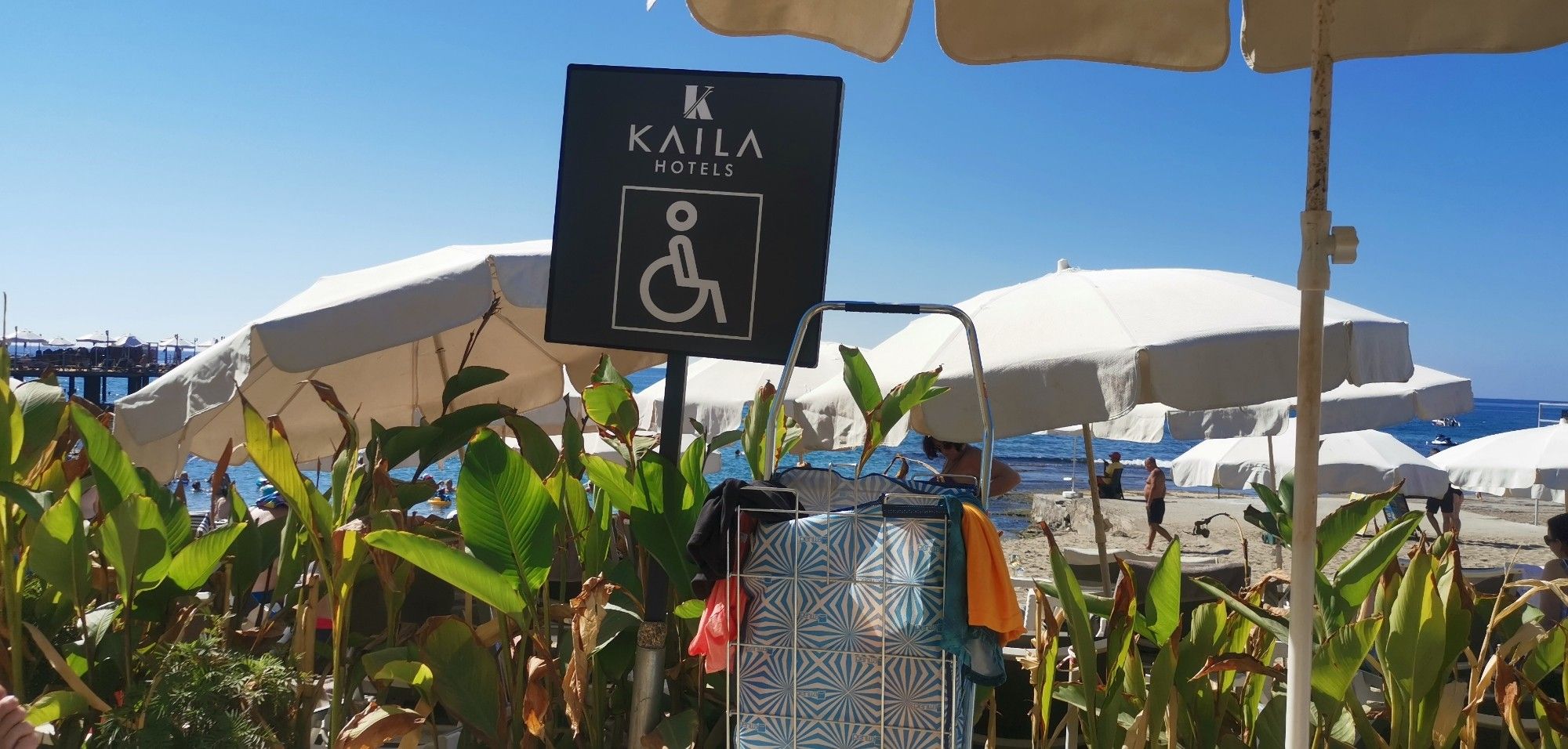 Bild vom Strand im Hintergrund mit Sonnenschirme. Eine Tafel mit einem Rollstuhl, das hier Behinderte Menschen einen guten Platz am Strand haben. Sehr barrierefrei.