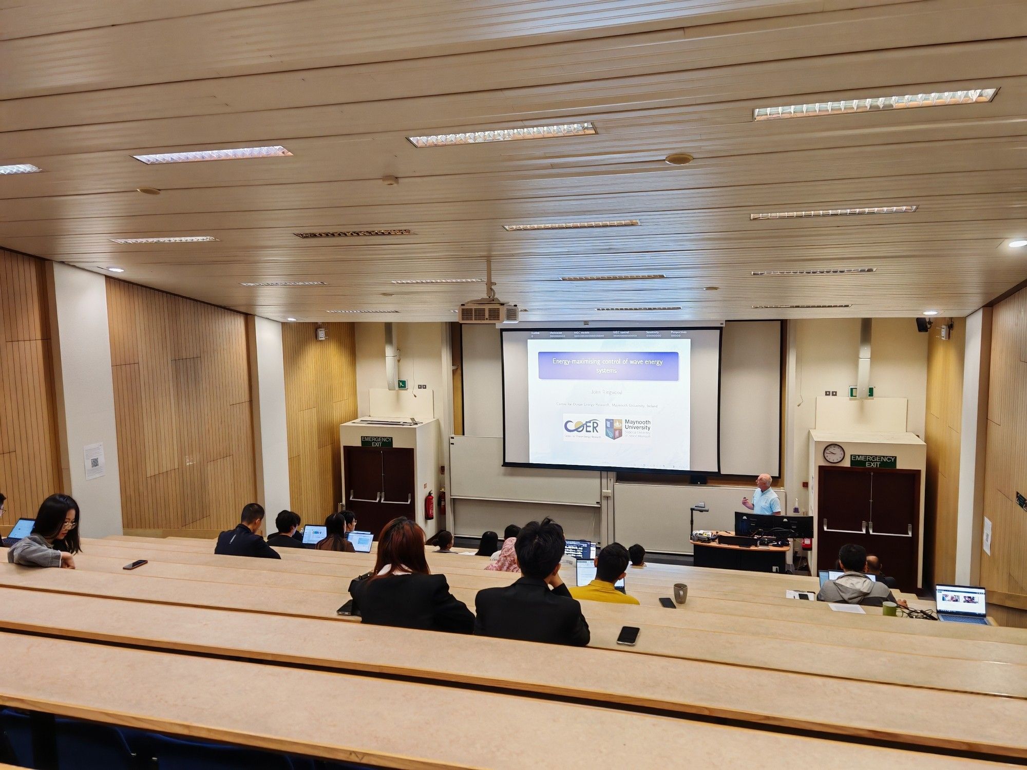 Prof John Ringwood delivering a talk in Rhodes Lecture Theatre while several students and colleagues altogether about 23 of them who are sitting around the lecture theatre attentively listening to the talk.