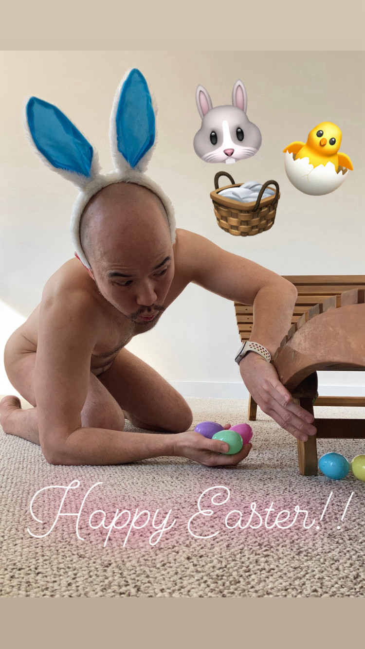 In naked man in bunny ears looking under chair and placing coloured eggs