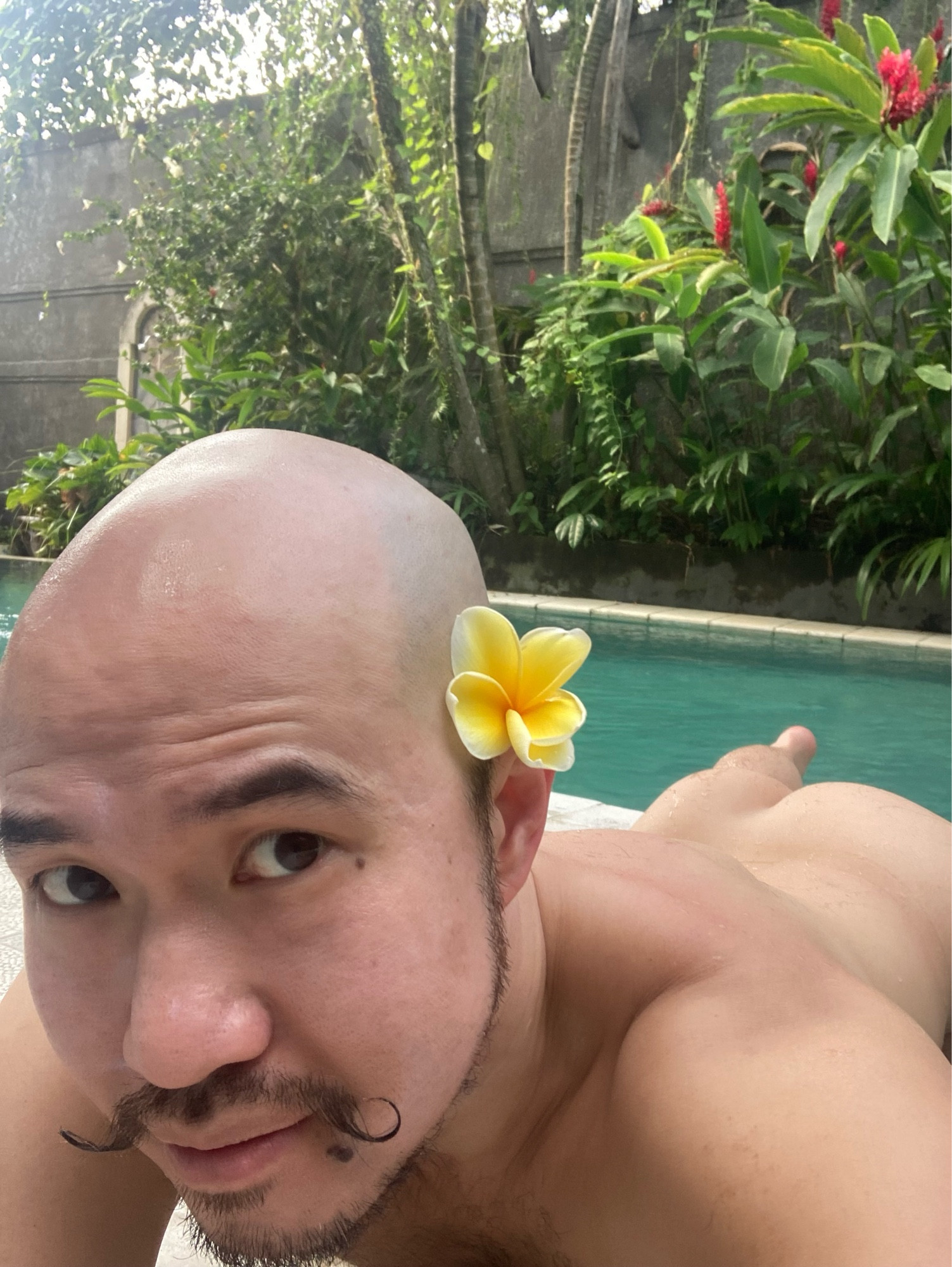 Naked mustache man lying next to a pool with yellow plumeria flower on one ear