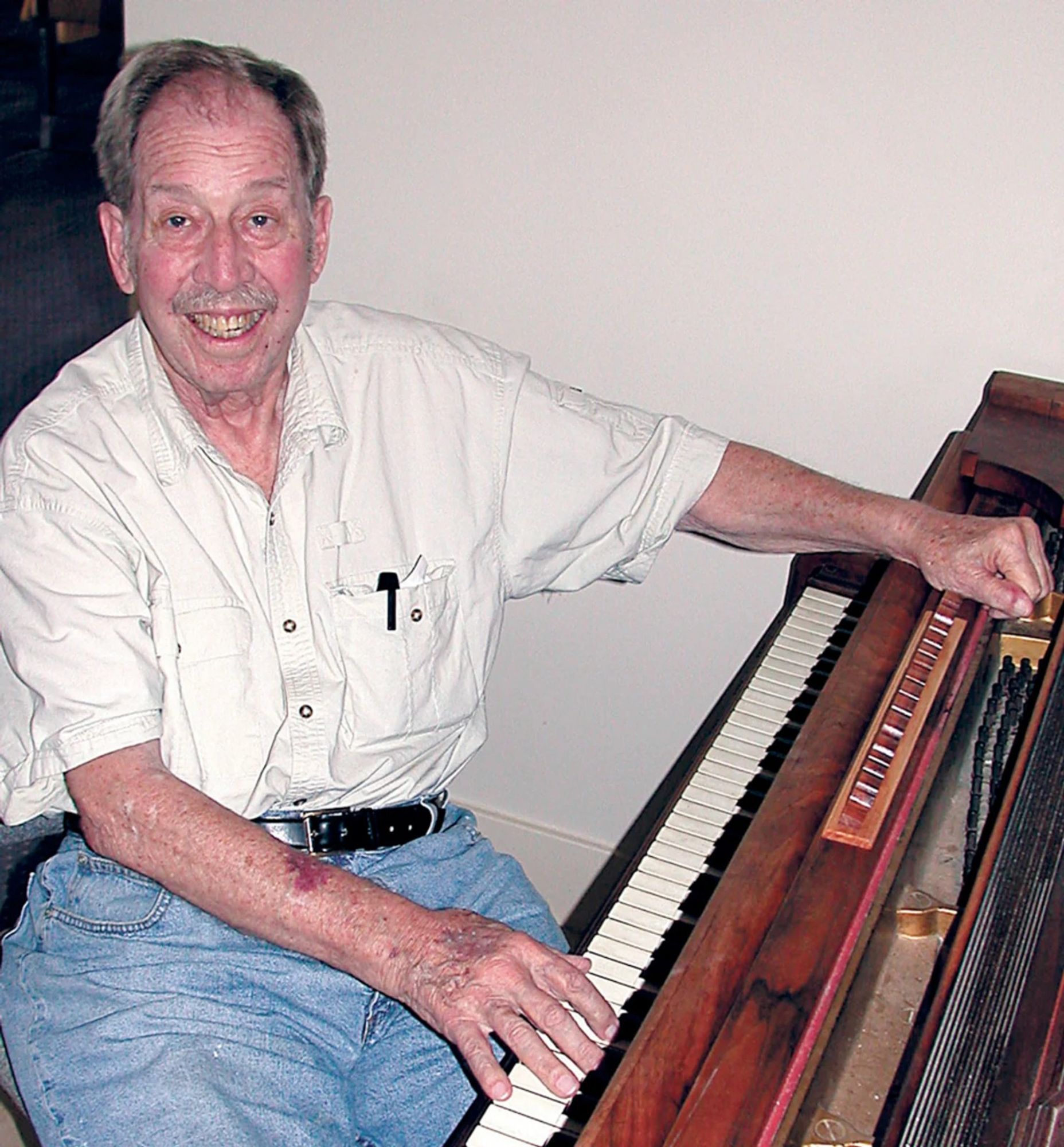 A imagem mostra Bob Bemer, o criador da tecla "Esc", sorrindo enquanto está sentado ao lado de um piano. Ele usa uma camisa clara de manga curta, com uma caneta no bolso, e calças jeans. Sua mão direita está sobre as teclas do piano.