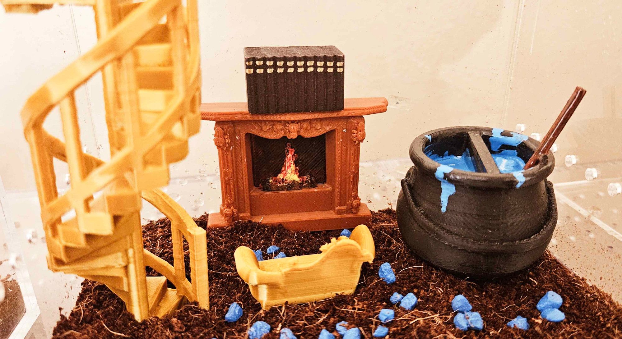 An acrylic jumping spider enclosure with soil substrate, small blue pebbles, a golden spiral staircase, a golden couch, a fake fireplace with tiny books on the mantle, and a cauldron with a blue potion inside