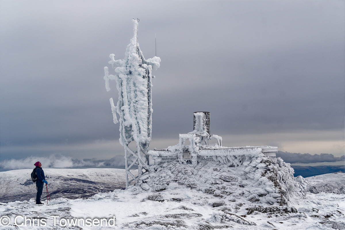 A weather station plastered with snow and ice on a mountain top with a hiker looking up at it and a cloudy sky