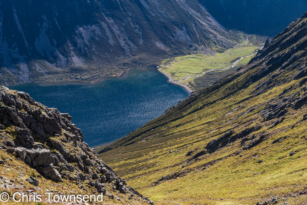 View down a steep rocky mountainside to a blue lake and a green meadow