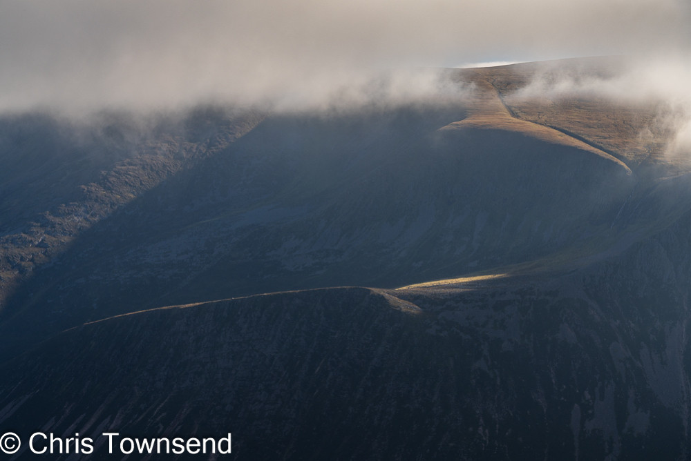 A dark cloud-capped mountainside with small areas lit by the sun