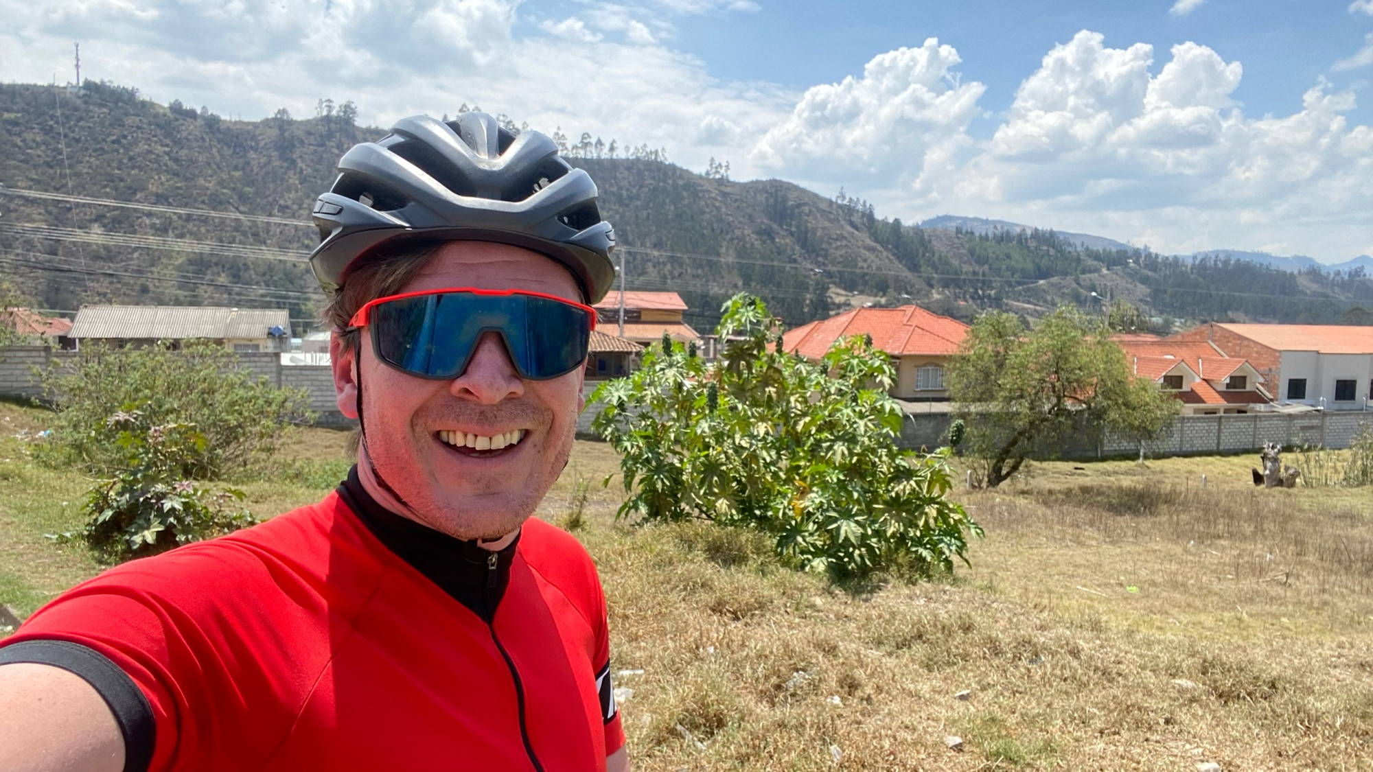 me smiling in my red kit and sunglasses, and black helmet. there’s a few houses and mountains behind me