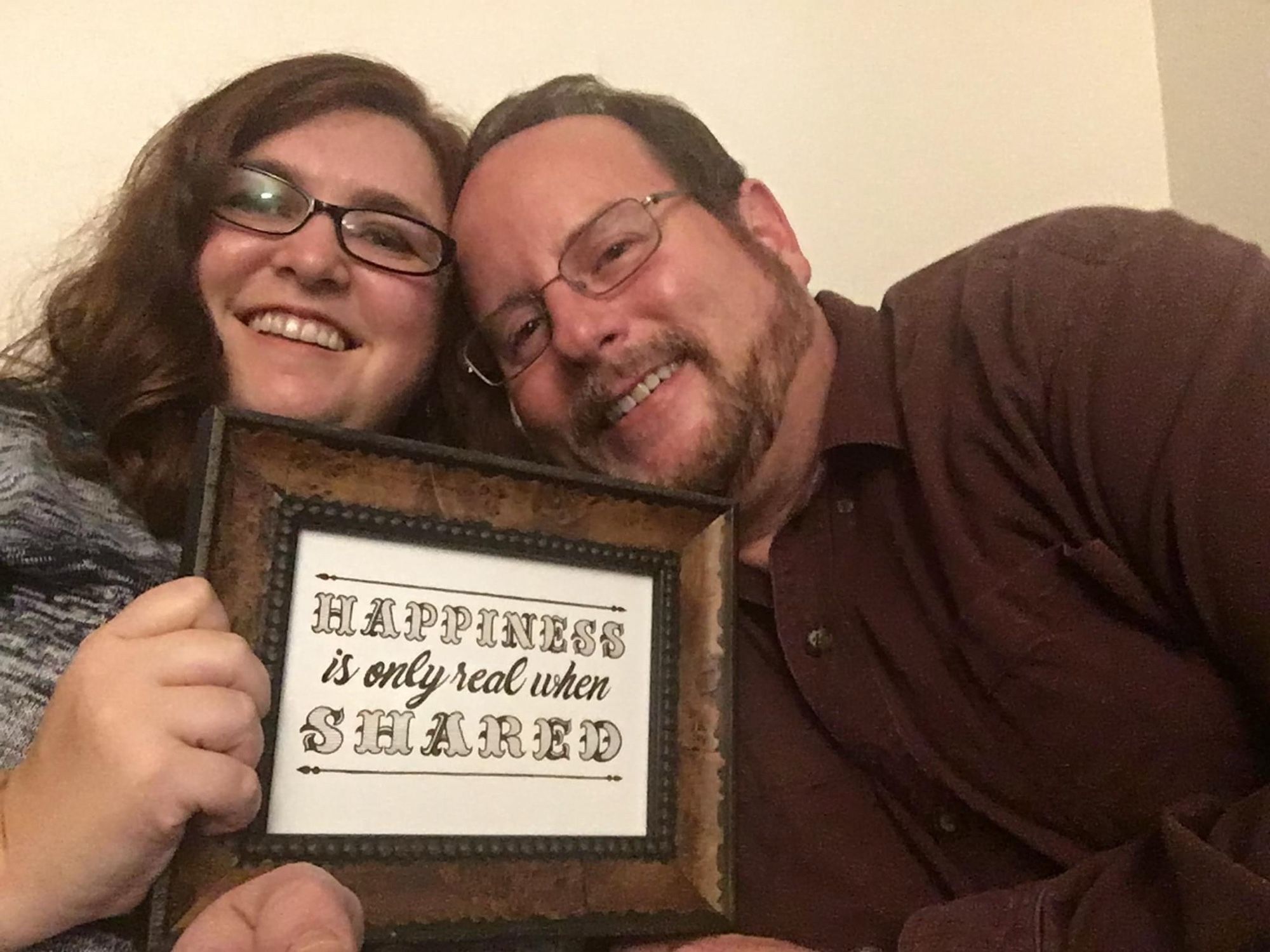 Me and my husband holding a framed calligraphy work by our friend Cougrr that says “Happiness is only real when shared”, a quote from Into the Wild