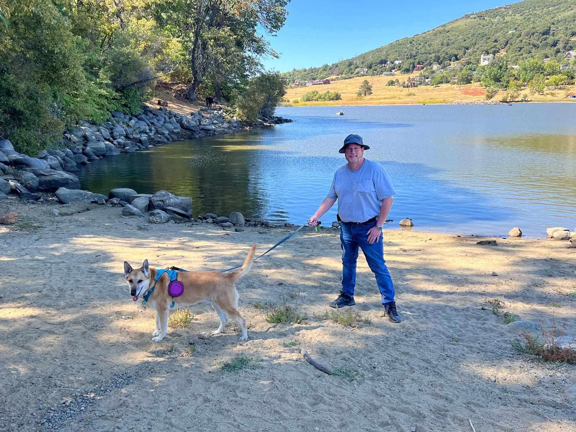 A bearded man, my husband, stands on the lakeshore with an all tan German shepherd mix wearing a teal harness and a purple collapsible water bowl attached to it. Lake is in the background, dog is to the left of the frame, man is in the center