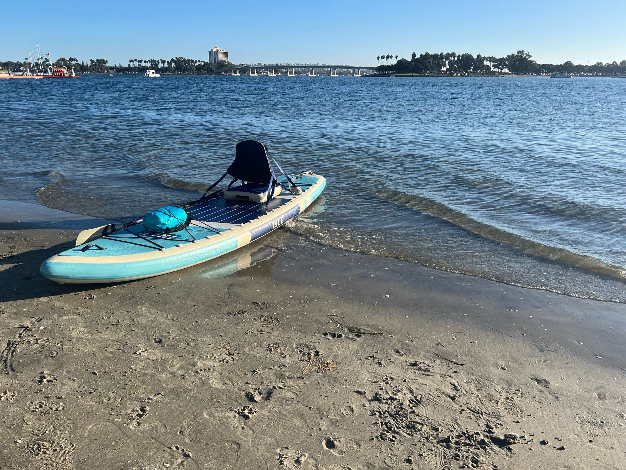 My teal ISLE Switch paddle board beached against a mission bay backdrop with a low bridge in the background