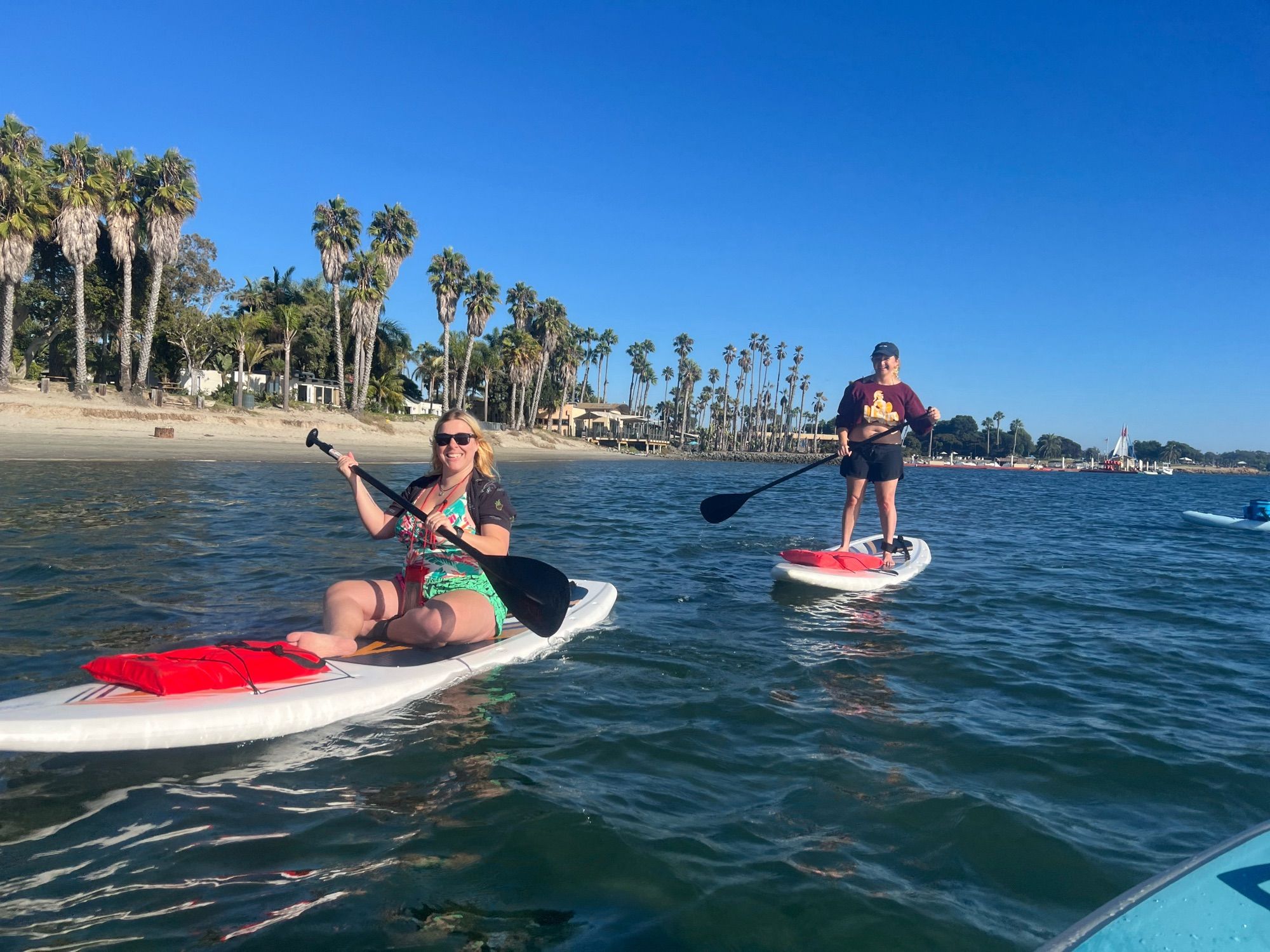 Two women on white paddle boards. One is sitting down, the other is standing up. They are facing me and smiling, the shore is to the left of the frame and there are palm trees