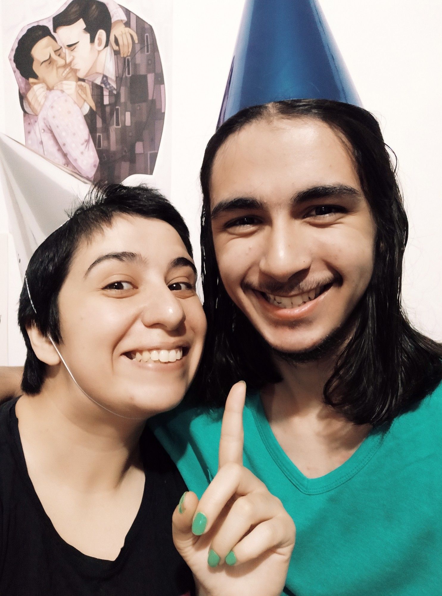 Uma foto minha sorrindo usando camiseta preta, cabelos curtos preto e chapéu de festa prata, apontando para o meu irmão que também sorri e está usando camiseta verde, cabelos longos pretos e um chapéu de festa azul.