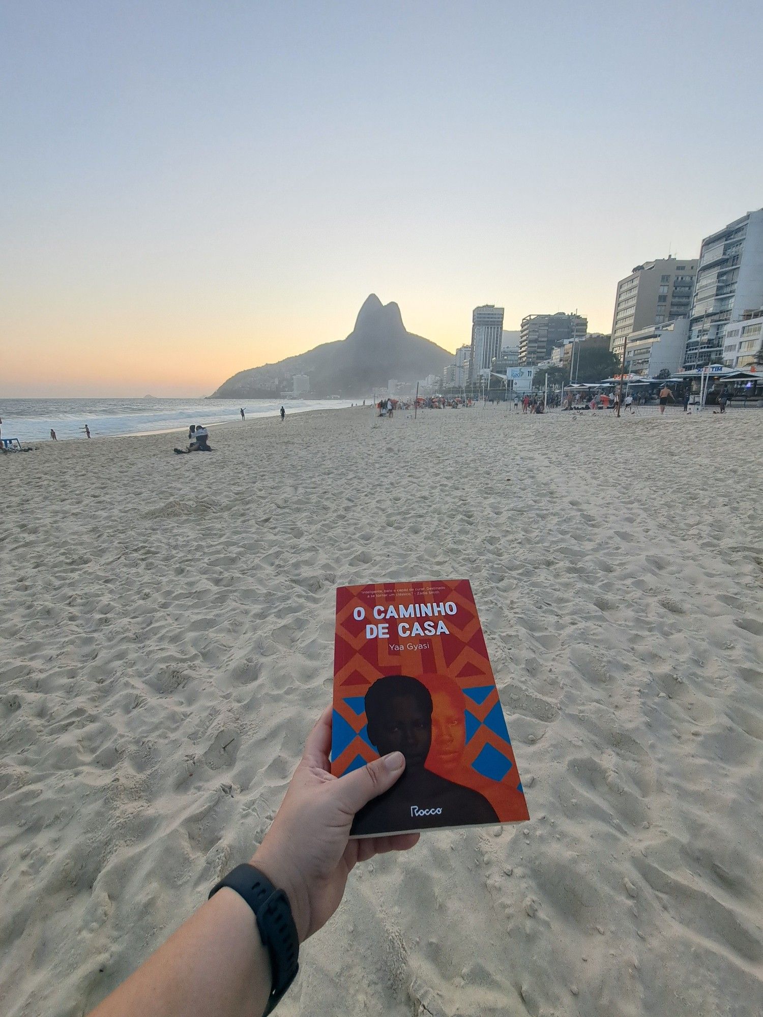 Mão com relógio de pulso segura livro de capa vermelha e laranja com duas mulheres negras na capa, uma delas está a frente e é retinta enquanto a outra é ela mesma mas alaranjada na parte de trás. O livro se chama O Caminho de Casa, da escritora ganesa Yaa Gyasi. Ao fundo da foto aparece a areia da praia e a praia do Leblon