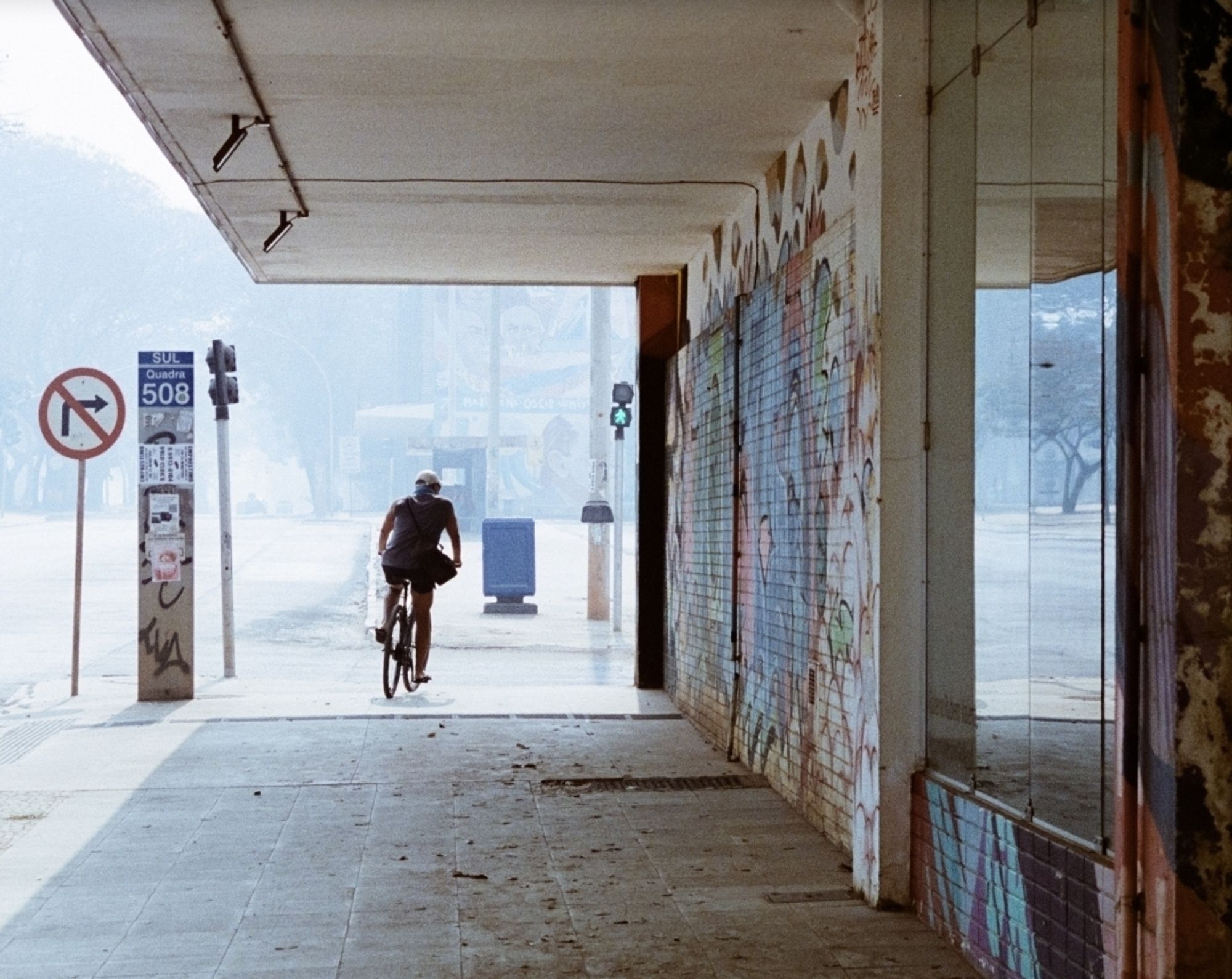 Silhueta de ciclista passando abaixo da marquise de um edifício com graffiti e, ao fundo, a rua esbranquiçada pela fumaça.